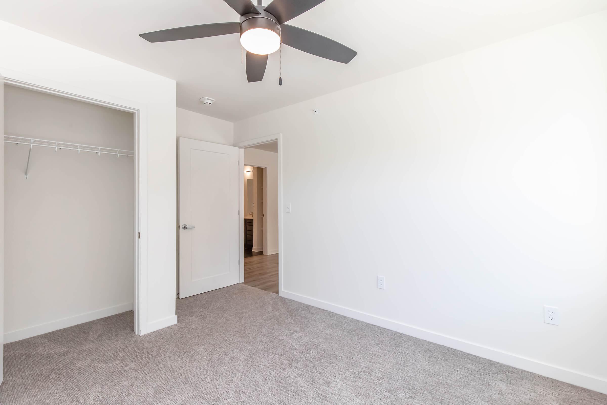 A bright and spacious room featuring a light gray carpet, a ceiling fan with a light fixture, and a single door leading to another area. There’s a closet with a hanging rod on the left, and a white wall with minimal decor, creating a clean and modern aesthetic.