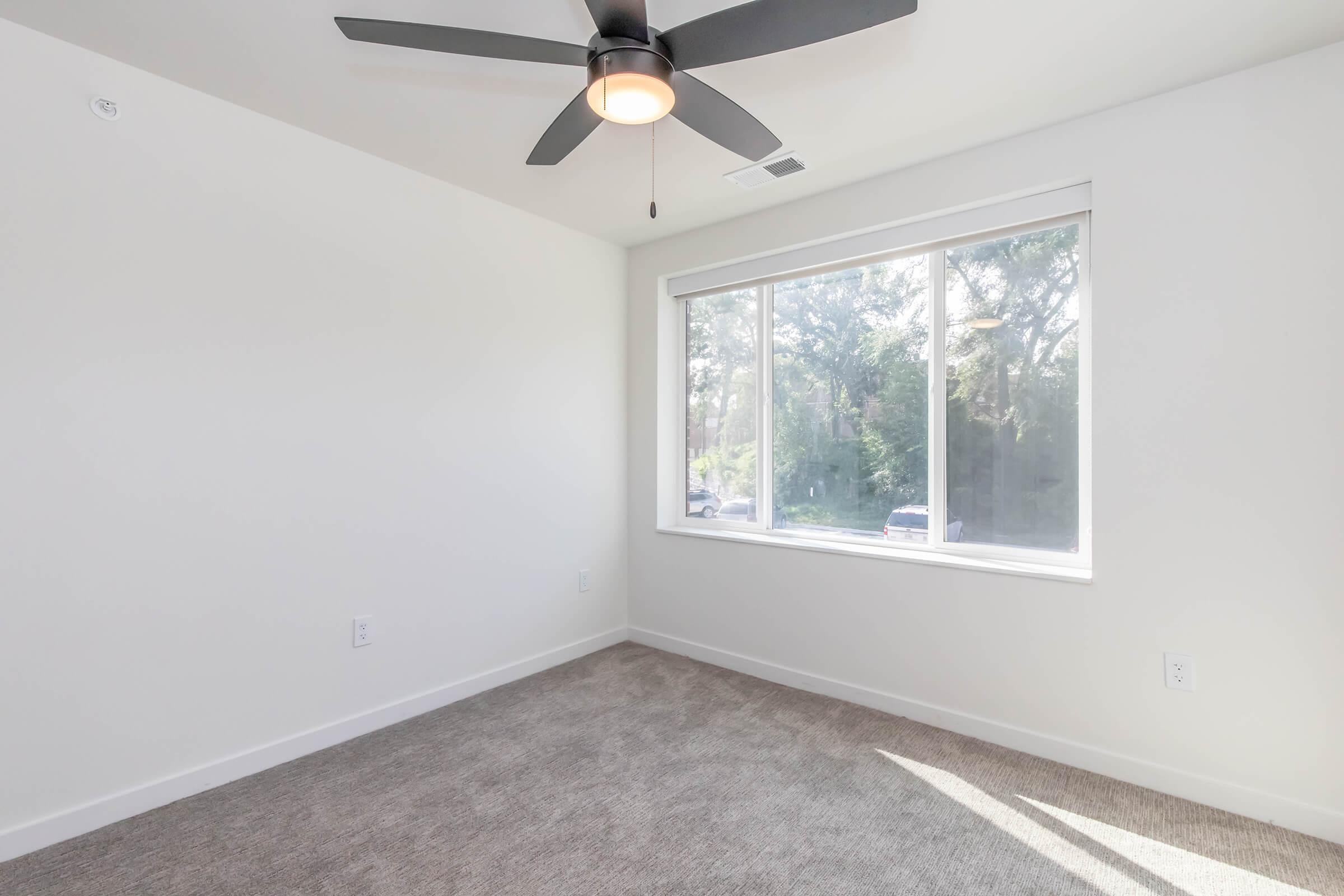 A well-lit, empty room featuring light gray walls and a plush carpet. A large window allows natural light to flood the space, revealing views of greenery outside. The ceiling is equipped with a modern ceiling fan, enhancing the room's contemporary design.