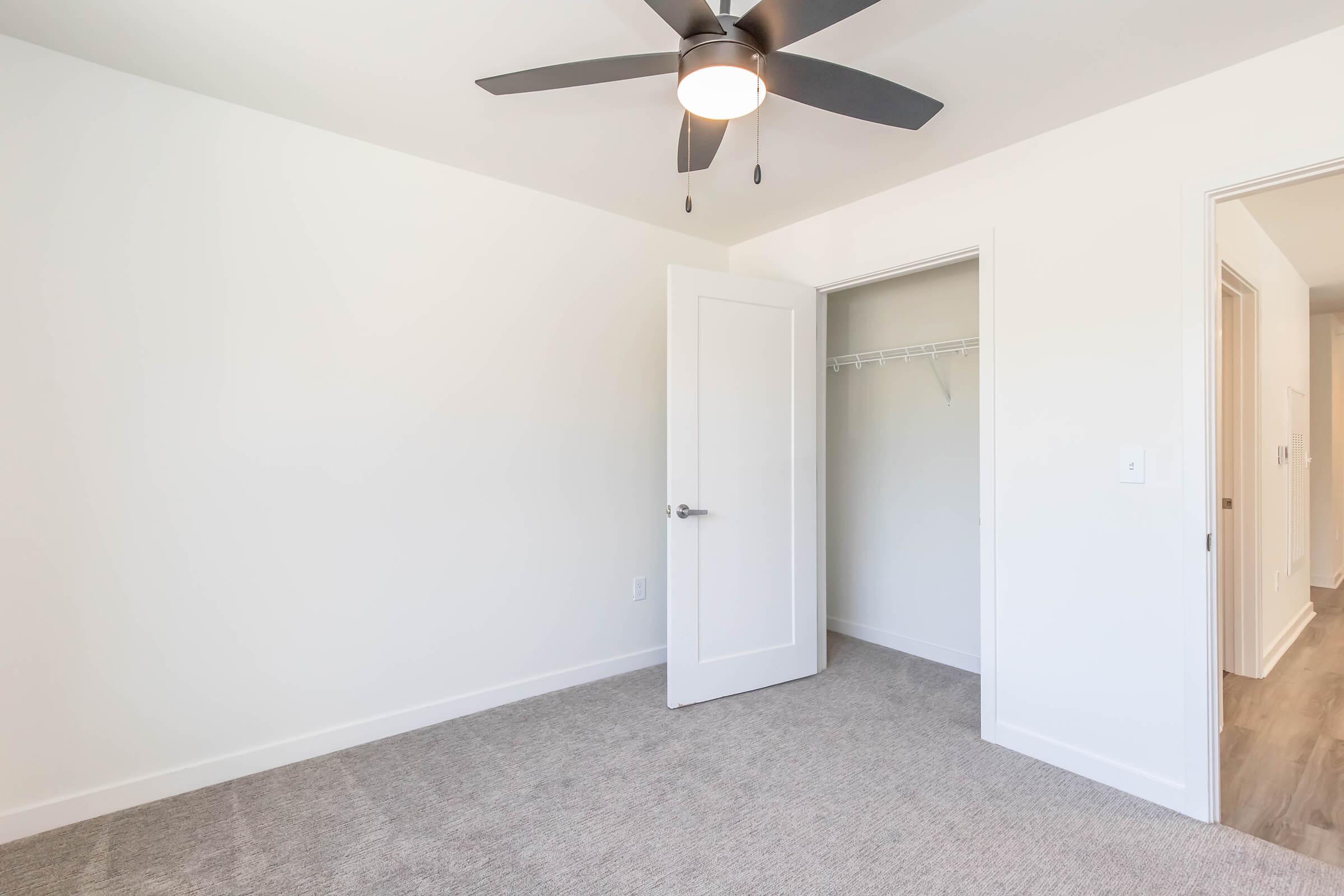 Empty room featuring gray carpet, a ceiling fan with light, and white walls. An open white door leads to a closet with a hanging rod and no shelves. The room is well-lit and minimalistic, suggesting potential for personalization.