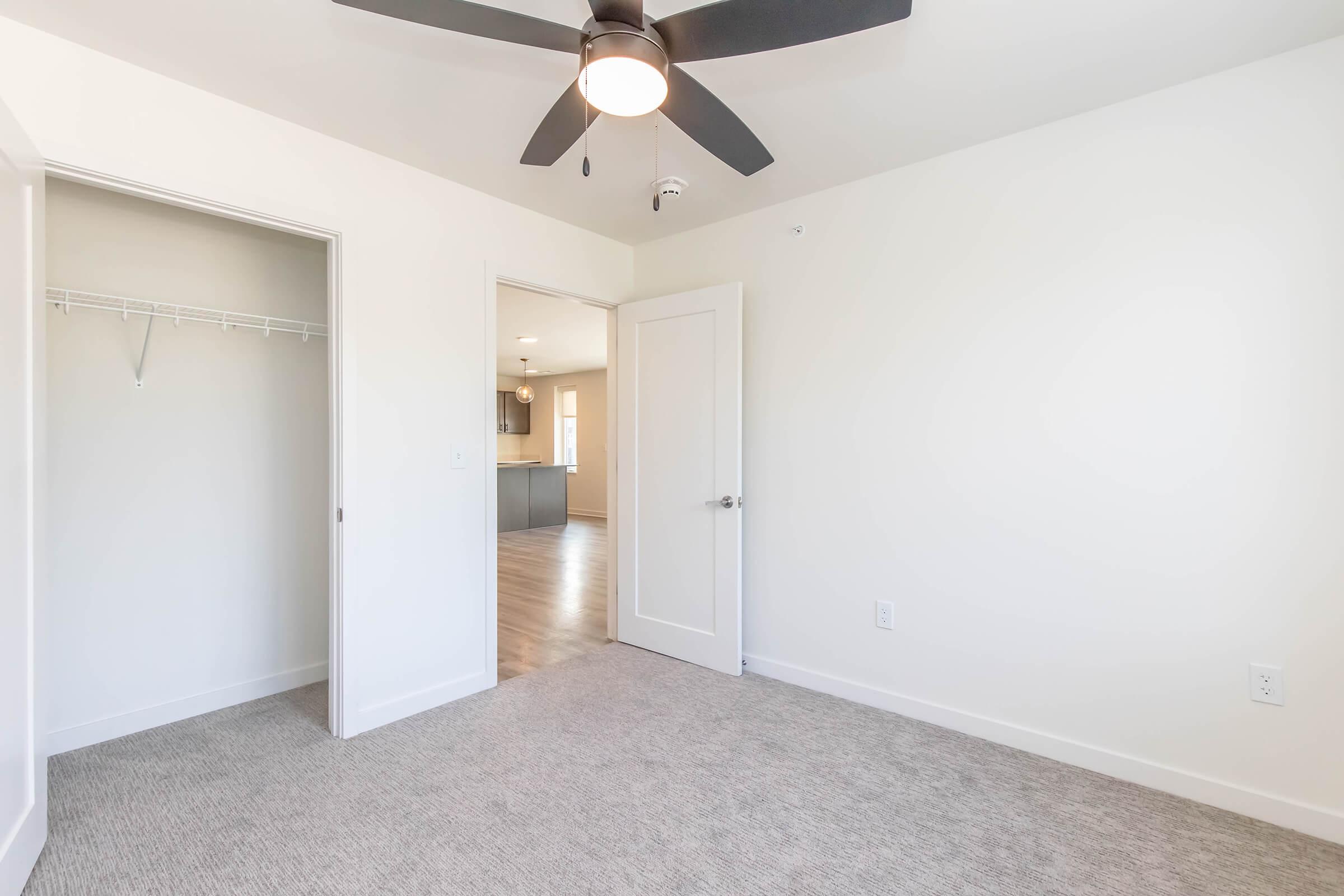 Empty room featuring light-colored walls, a ceiling fan, and beige carpet. The room has a door leading to a kitchen area visible in the background and a closet with a single rack on one side. Natural light comes through the doorway, creating a bright and airy atmosphere.