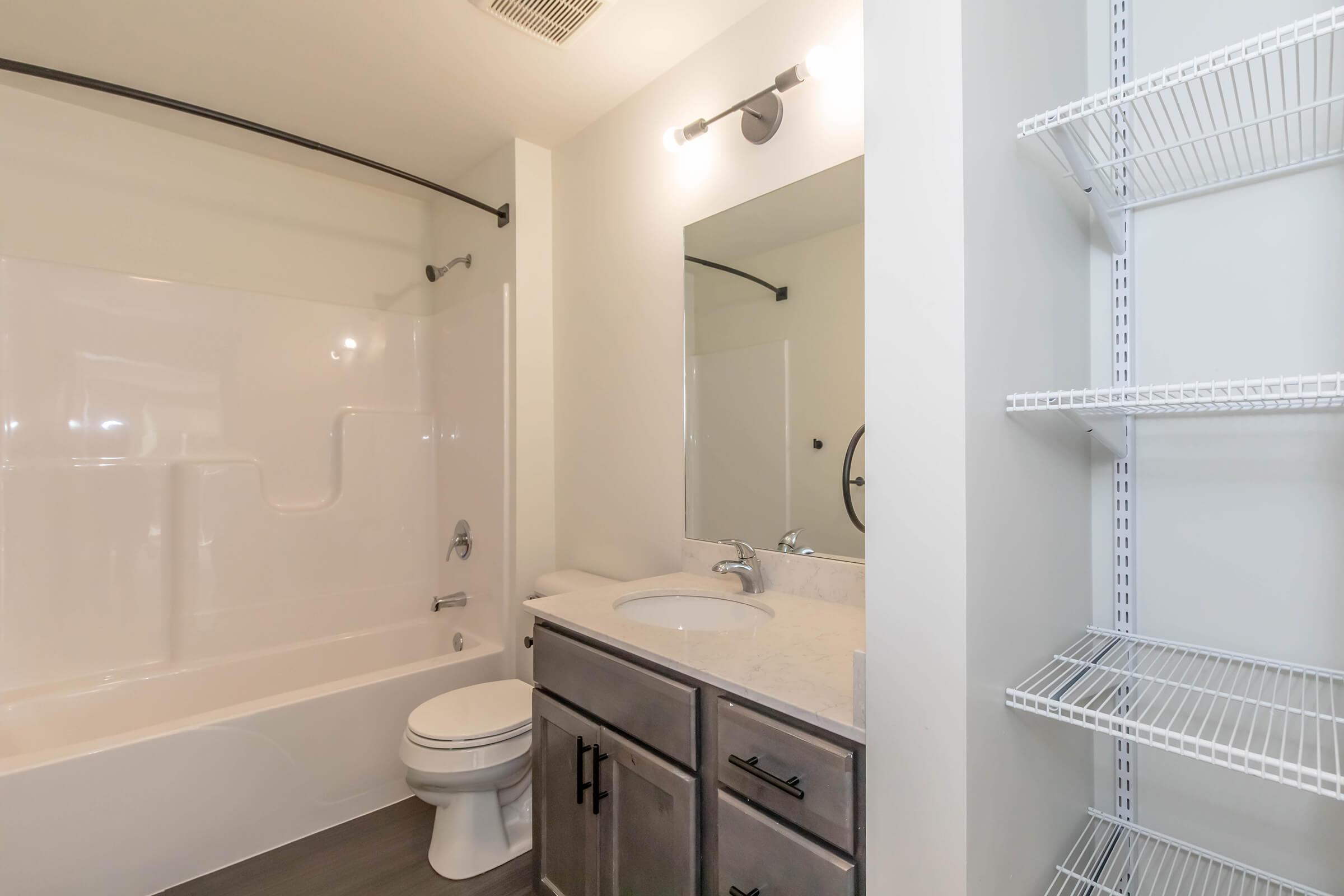 A modern bathroom featuring a white tub and shower, a sleek vanity with a light-colored countertop, a round mirror, and a small shelving unit on the right for storage. The walls and floor have neutral tones, contributing to a clean and spacious look.