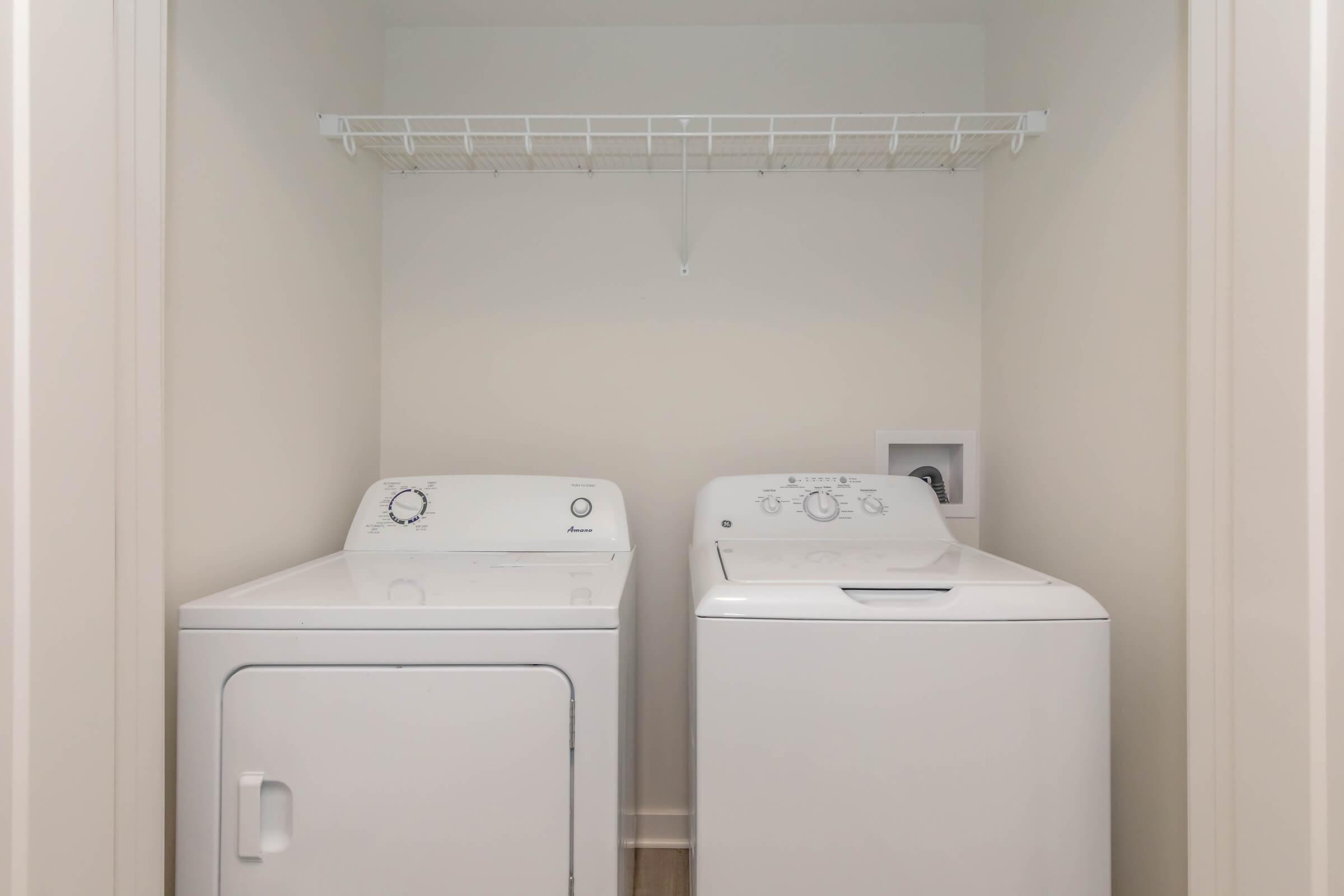 A clean laundry room featuring a white top-loading washing machine and a white dryer side by side. Above them, there is a wire shelf for storage, set against a neutral-colored wall. The floor is wooden, adding a cozy touch to the space.