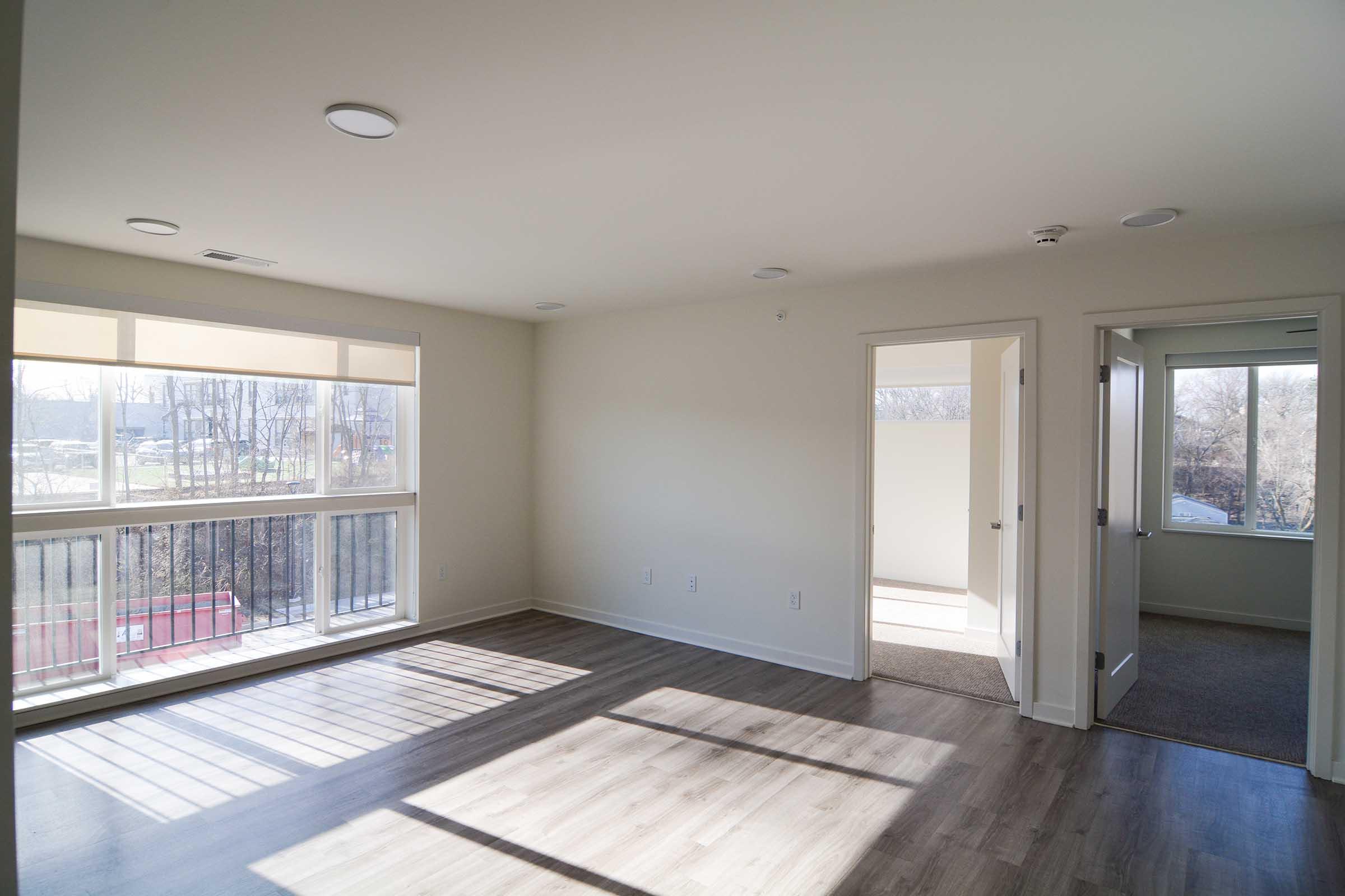 A bright, empty room with light-colored walls and large windows letting in natural light. The space features hardwood flooring, with a doorway leading to another room. In the background, tree branches are visible through the windows, suggesting a serene outdoor view.