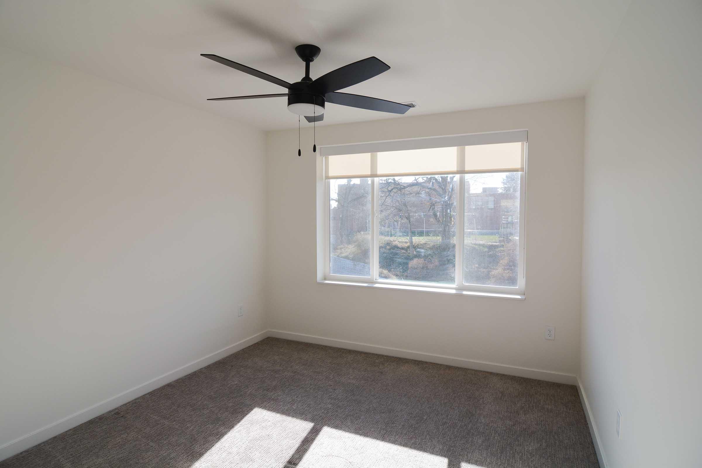 A bright, empty room with a ceiling fan and a large window letting in natural light. The walls are painted white, and the floor is covered with light gray carpet. Outside the window, trees and greenery are visible, suggesting a peaceful outdoor view.