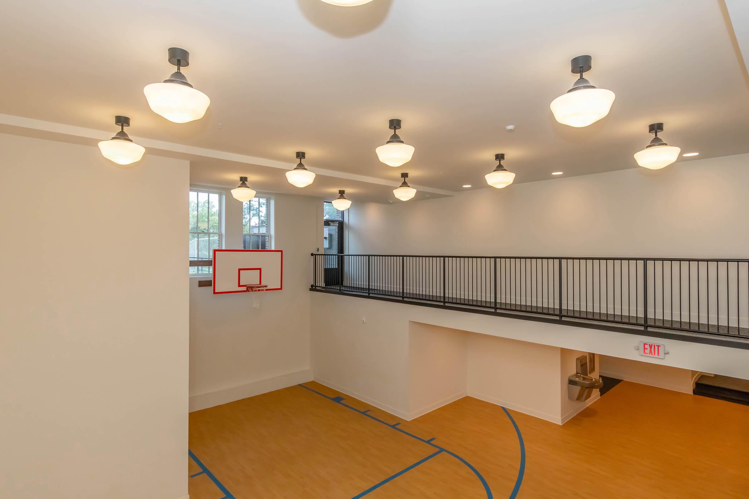 View of a modern indoor basketball court featuring a hoop mounted on a wall. The court has a wooden floor with blue lines marking the playing area. The space is well-lit with multiple ceiling fixtures and has an open upper level with a railing overlooking the court. An exit sign is visible on the wall.