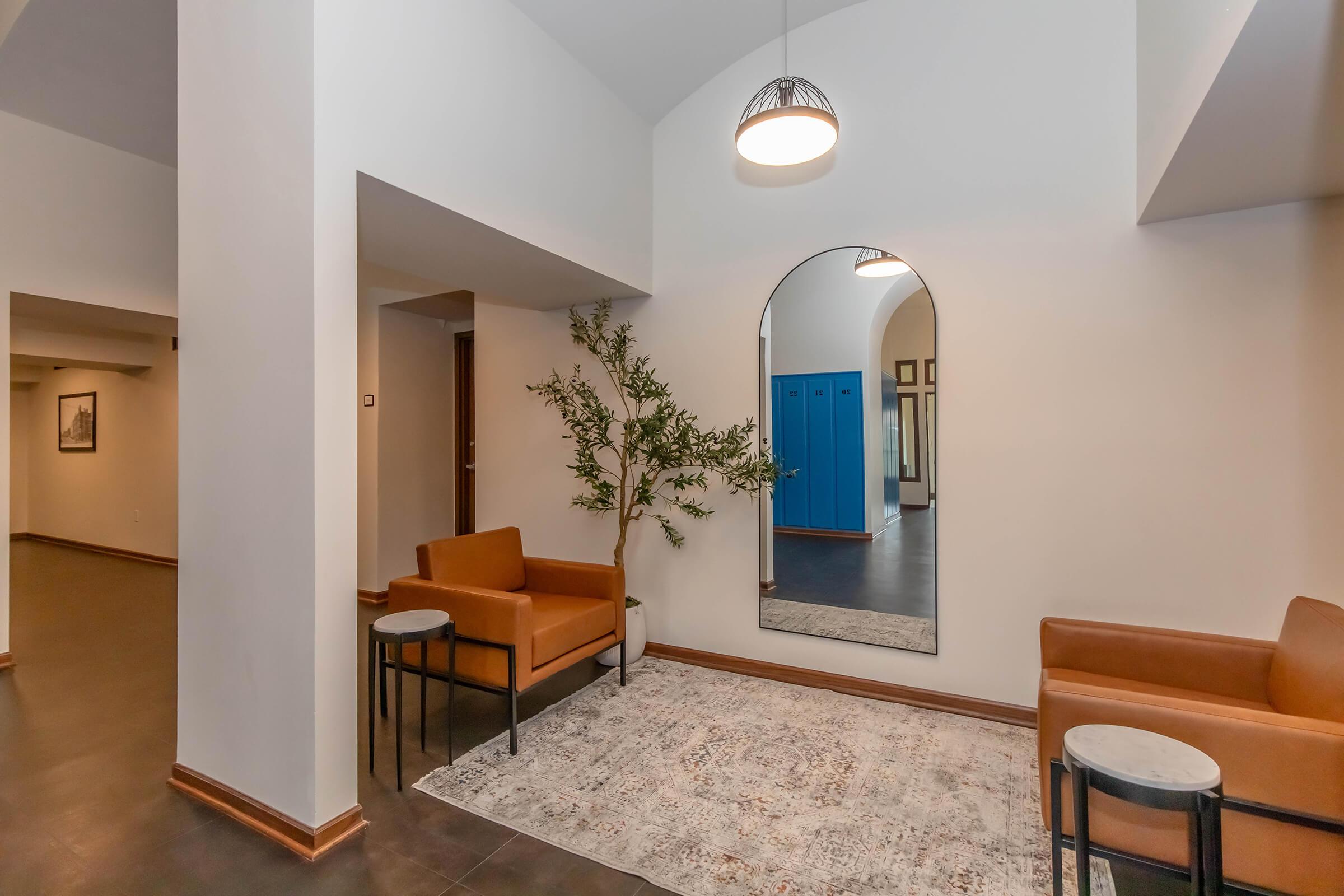 Modern interior space featuring a minimalist design with two brown chairs and small side tables. A large mirror is mounted on the wall adjacent to a decorative plant and a textured rug. In the background, there are blue lockers and an open area, creating a spacious and inviting atmosphere.