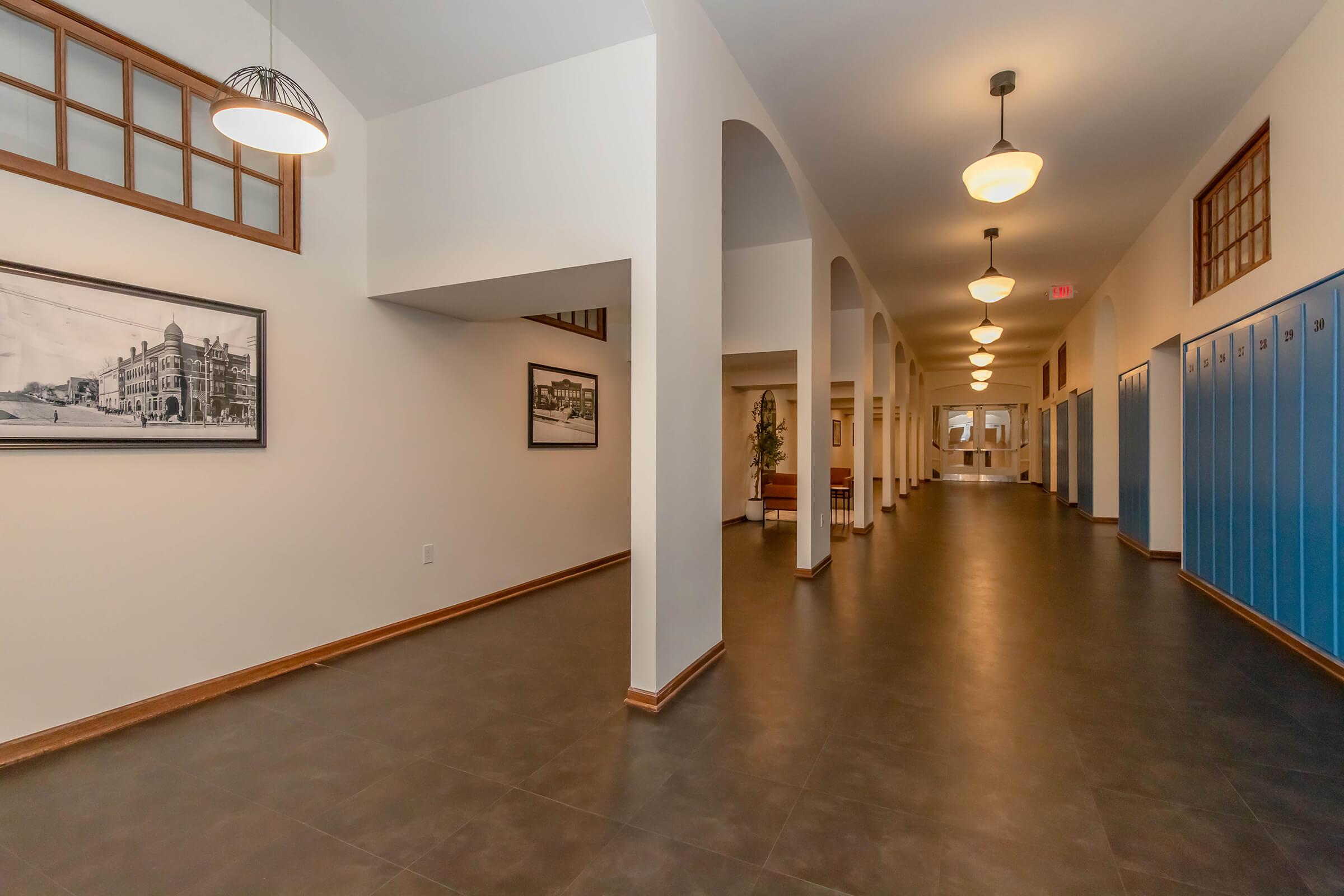 A well-lit school hallway featuring high ceilings, archways, and a series of blue lockers on one side. Black-and-white historical photographs adorn the walls, and pendant lights hang from the ceiling, creating an inviting atmosphere. The floor is dark tiles, enhancing the modern yet historic feel.