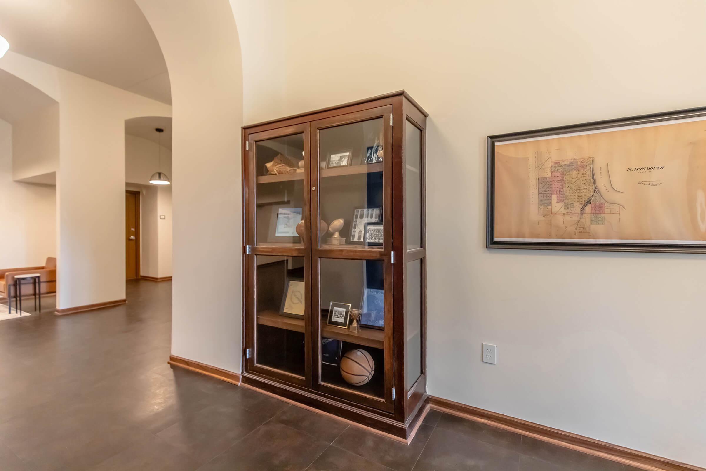 A wooden display cabinet with glass doors, containing various framed photographs and memorabilia. On the floor near the cabinet is a basketball. A framed map is mounted on the wall beside the cabinet, and the space features neutral-colored walls and a polished floor.