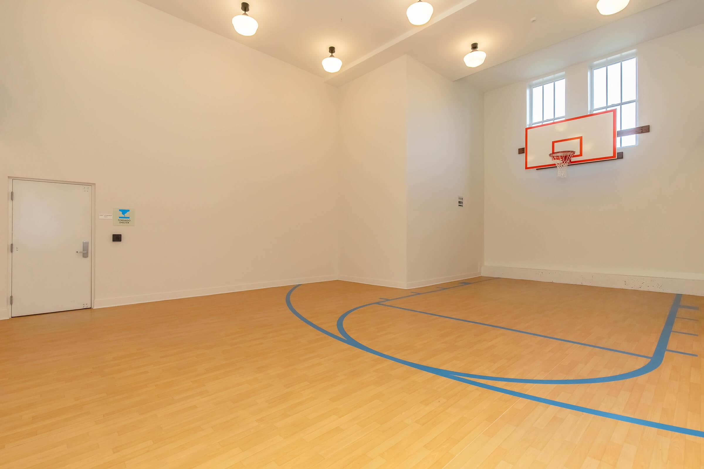 An indoor basketball court featuring a wooden floor with blue court lines, a basketball hoop mounted on the wall, and bright ceiling lights. The room is well-lit with tall windows, providing a spacious and inviting atmosphere for recreational activities.