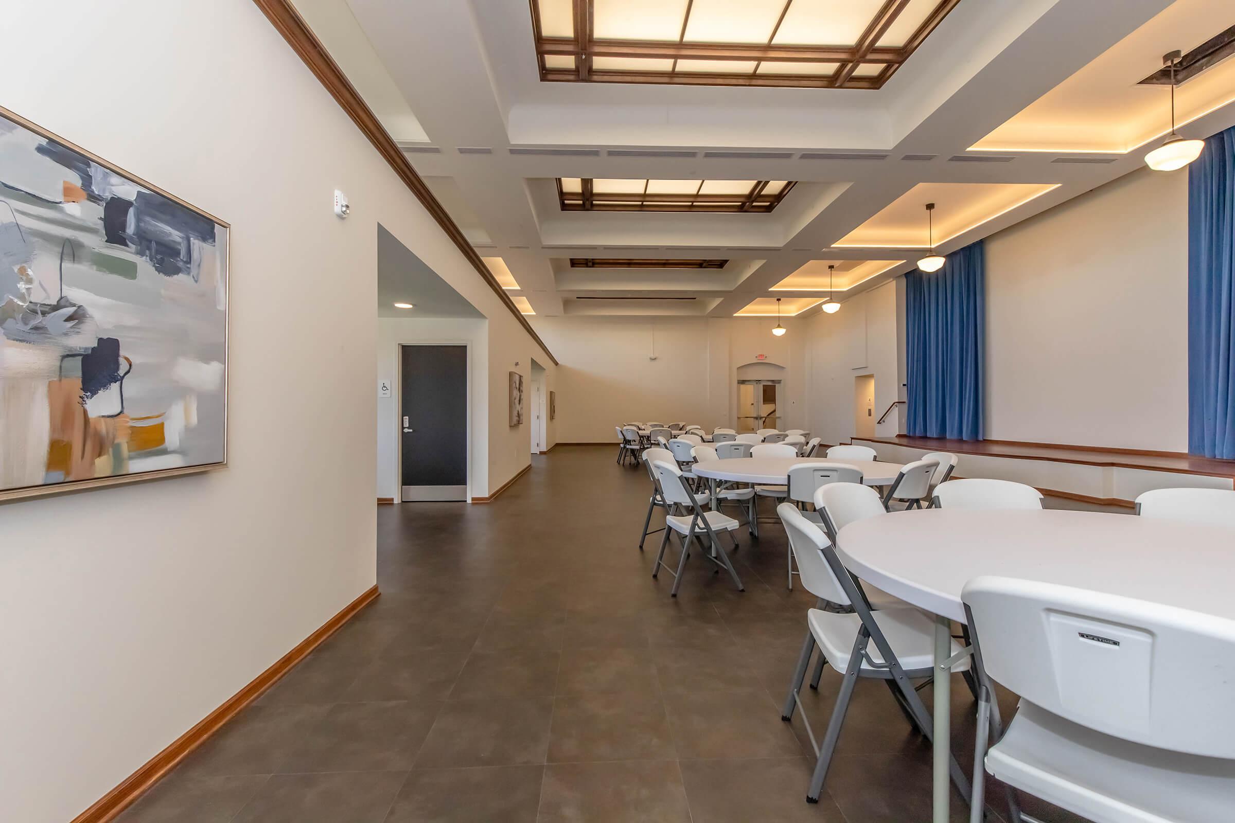 A spacious, well-lit room featuring several round tables with chairs. The walls are painted a soft color, and large windows allow natural light. Artwork is displayed on one wall, while blue curtains complement the neutral decor. The space appears suitable for gatherings or events.