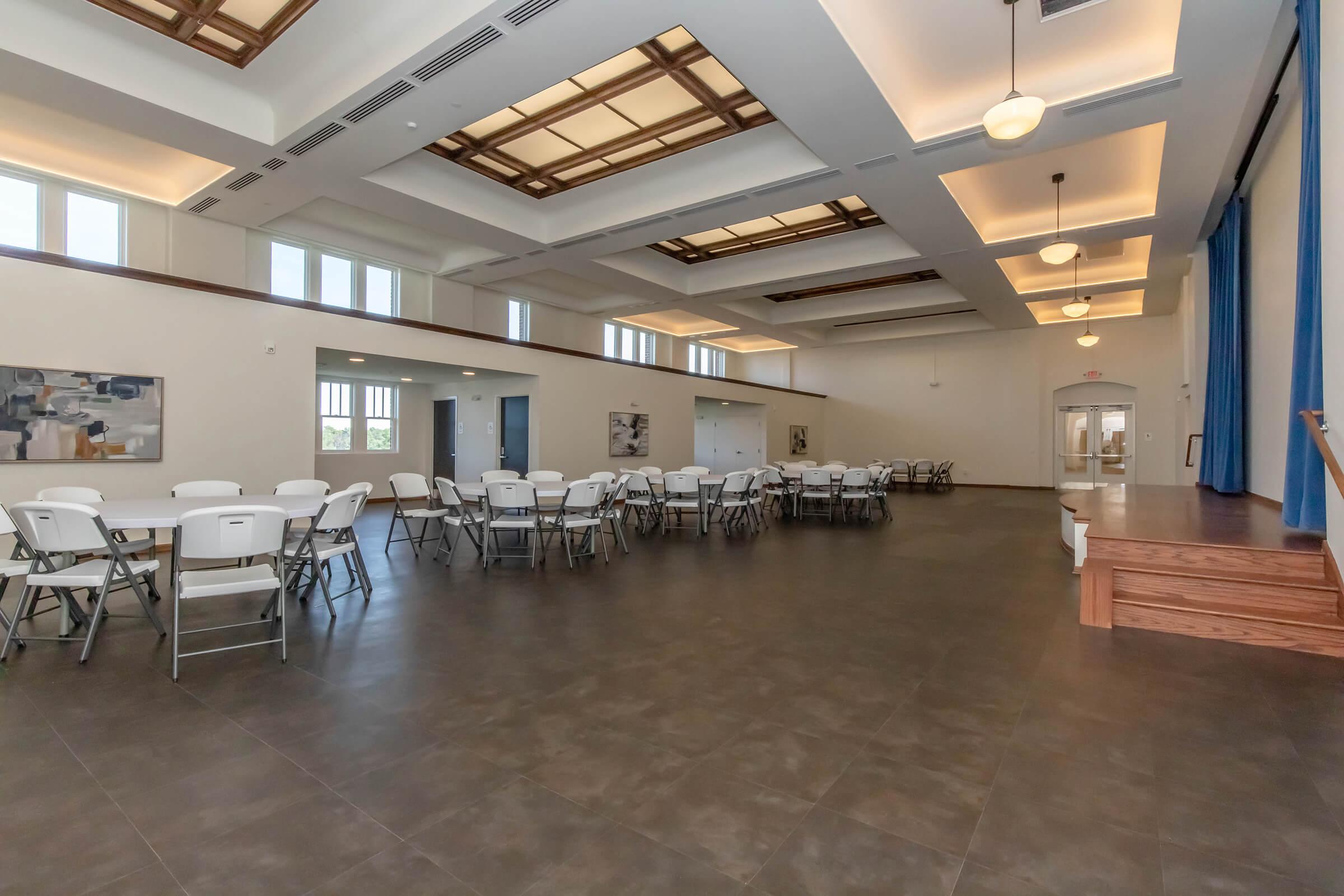 An empty indoor event space featuring several round tables with chairs arranged, a wooden stage area, and large windows letting in natural light. The ceiling has a decorative design, and the walls are painted in neutral colors, creating a spacious and inviting atmosphere.