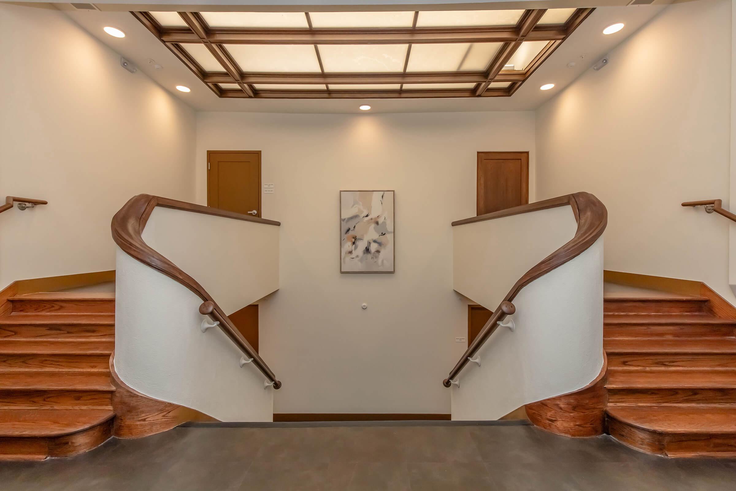 Elegant staircase featuring two sweeping wooden railings, leading to a well-lit landing. Soft neutral walls and floors enhance the modern aesthetic, complemented by a decorative painting on the wall. A glass skylight allows natural light to illuminate the space, creating a warm and inviting atmosphere.