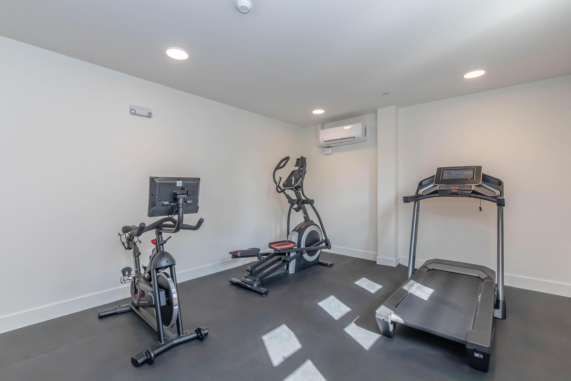 A modern gym interior featuring exercise equipment including an elliptical machine, a stationary bike, and a treadmill. The room has white walls, bright overhead lighting, and an air conditioning unit, providing a clean and well-lit environment for workouts.