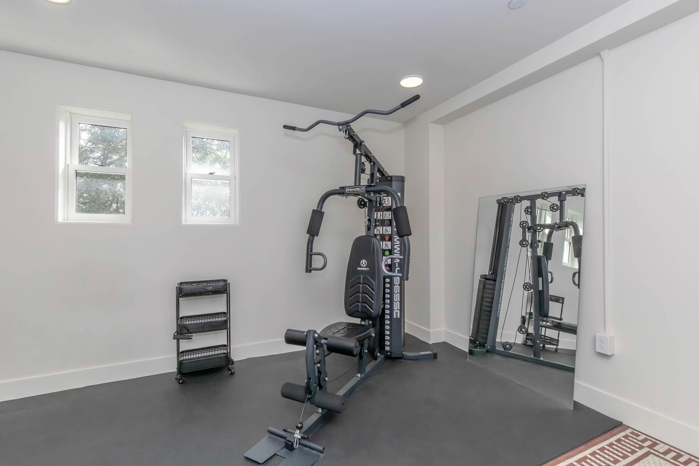 A home gym featuring a multi-gym workout machine, a mirror, and a black cart for equipment storage. The room has white walls and two small windows, providing natural light. The flooring is dark, creating a modern and functional workout space.