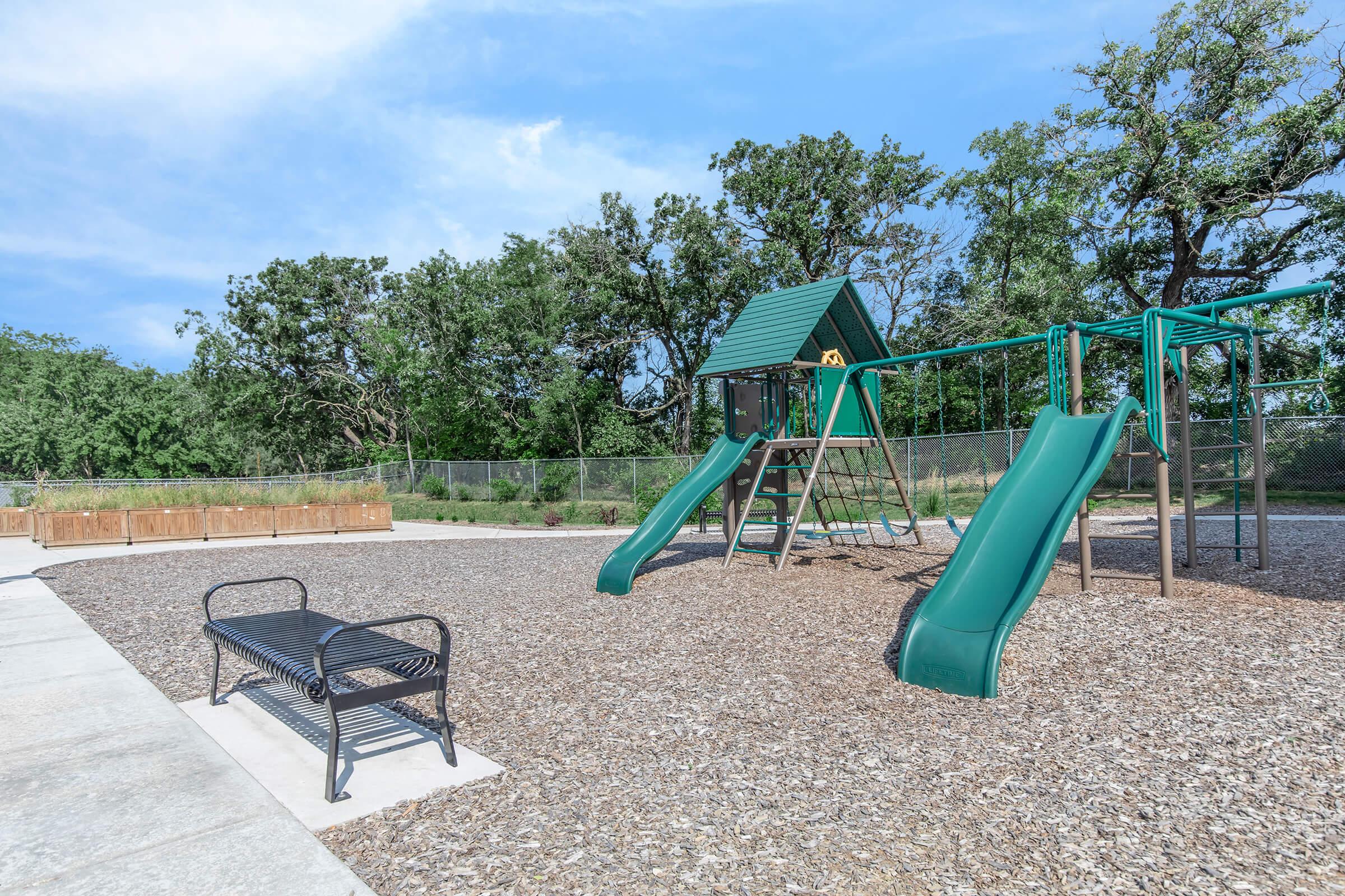 Playground scene featuring a green playset with two slides, a climbing structure, and a sandbox area. There is a black metal bench nearby, set on a gravel surface, surrounded by trees and a fenced area in the background under a clear blue sky.