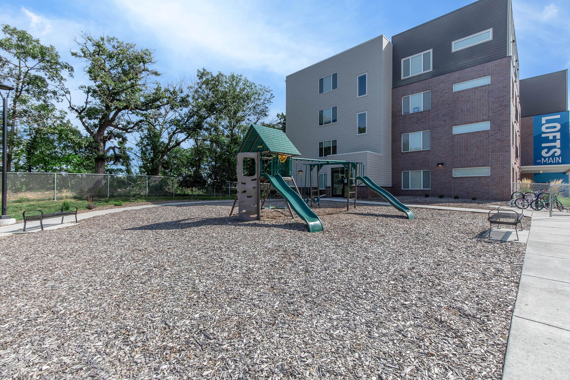 Playground area with a green slide structure and tan play equipment surrounded by wood chips. In the background, a modern apartment building with a sign reading "LOFTS" and "MAIN" is visible. Trees and fencing are present, creating a cozy outdoor space.