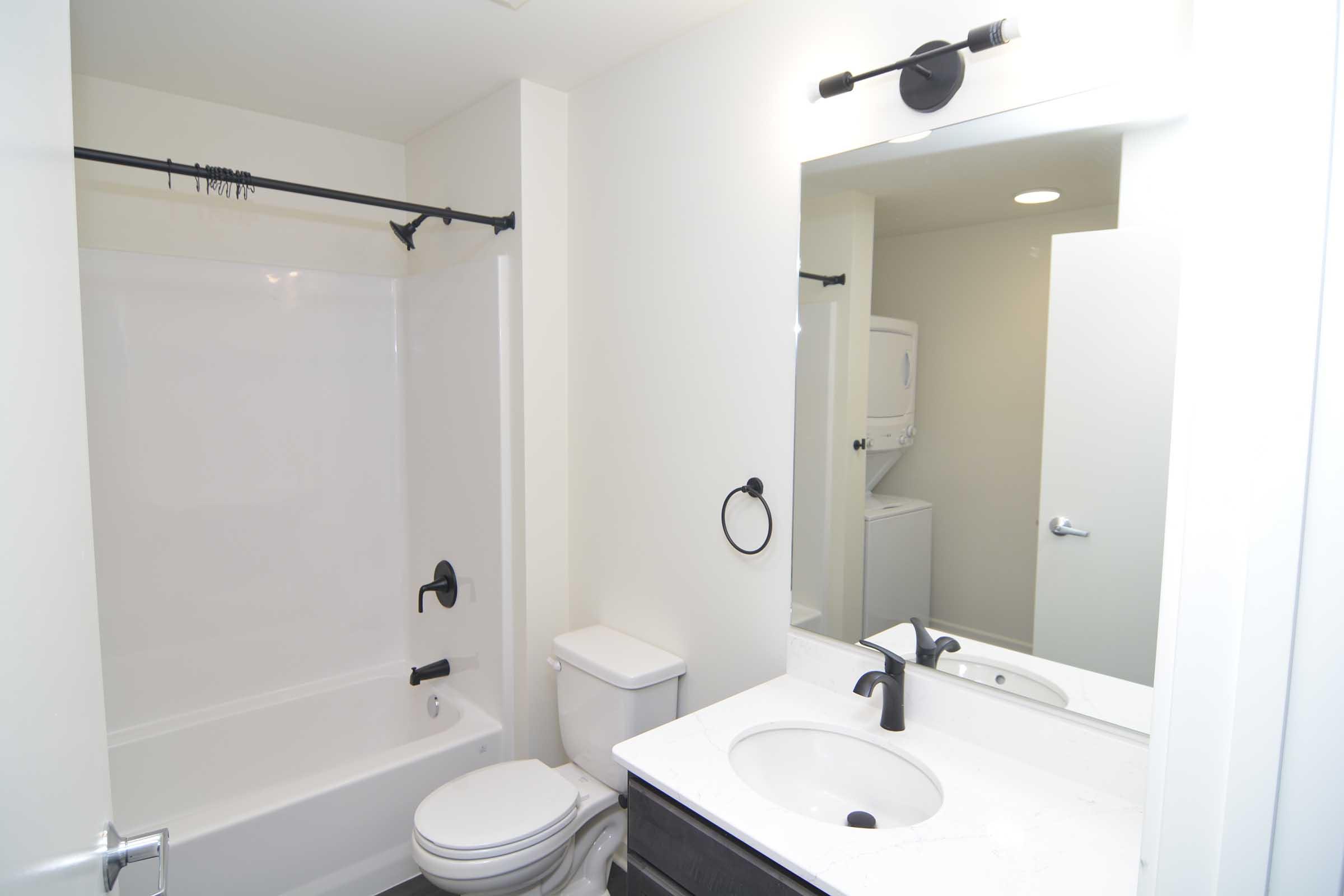A modern bathroom featuring a white bathtub with a black faucet, a toilet, and a sink with a black faucet. A large mirror reflects the space, and there's a washer and dryer unit visible in the corner. The walls are painted white, creating a bright and clean atmosphere.