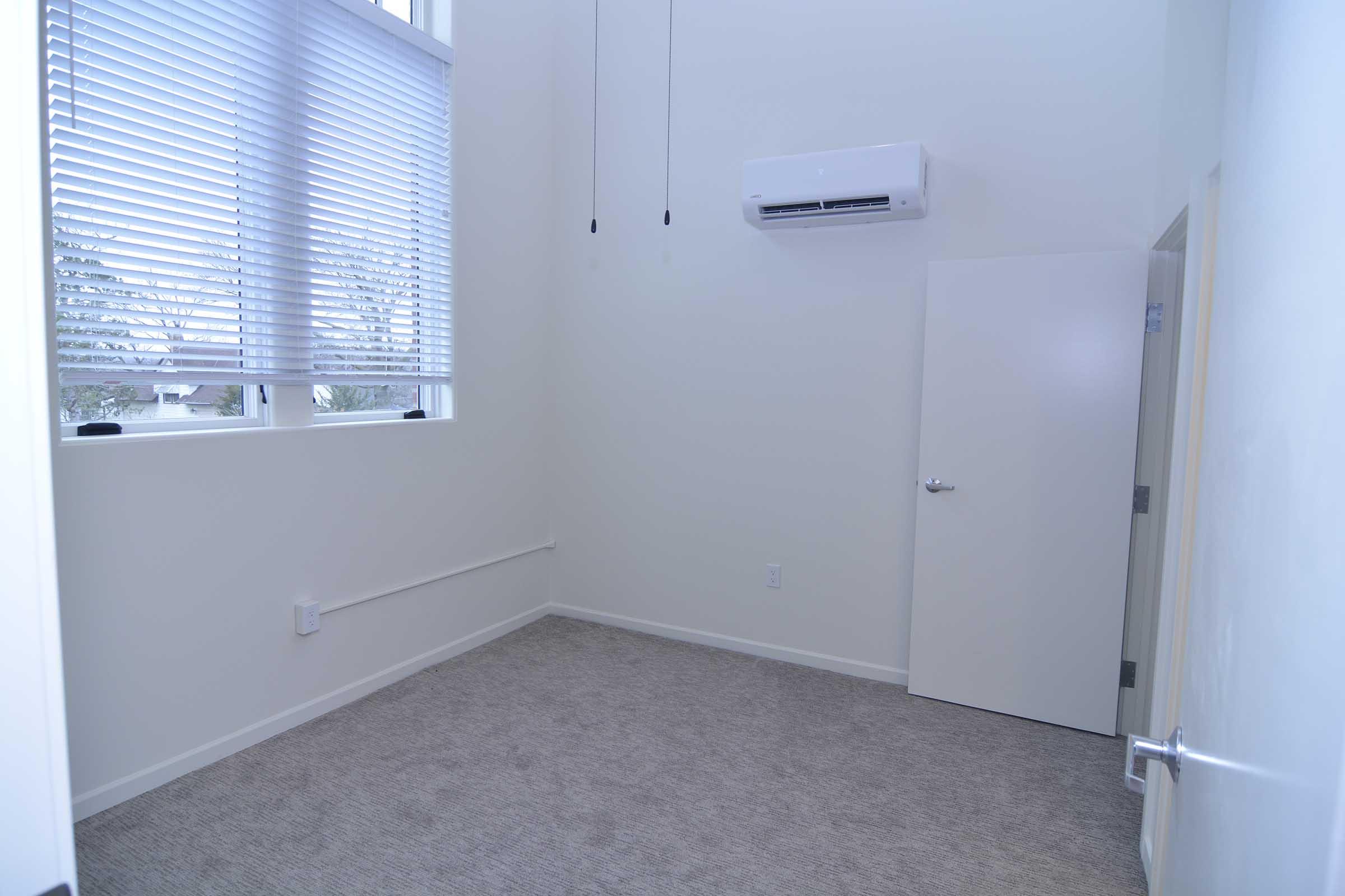 A well-lit, empty room featuring light-colored walls and carpet. It has large windows with blinds, an air conditioning unit on the wall, and a door leading to another space. The room has a minimalist design and is devoid of furnishings.