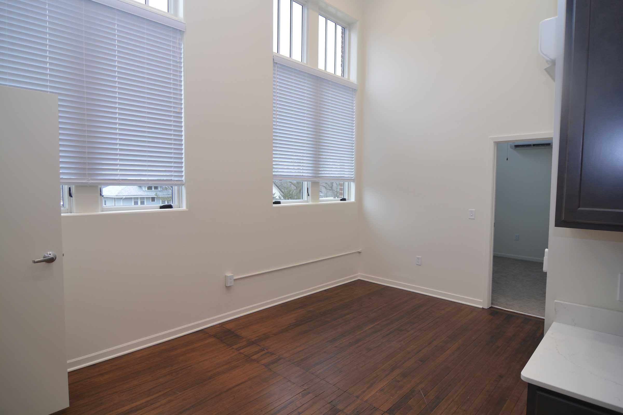 A well-lit room with high ceilings and large windows featuring white blinds. The floor is polished hardwood, and there is a plain white door on the left side. The walls are painted a light color, and the background shows an open doorway leading to another room.