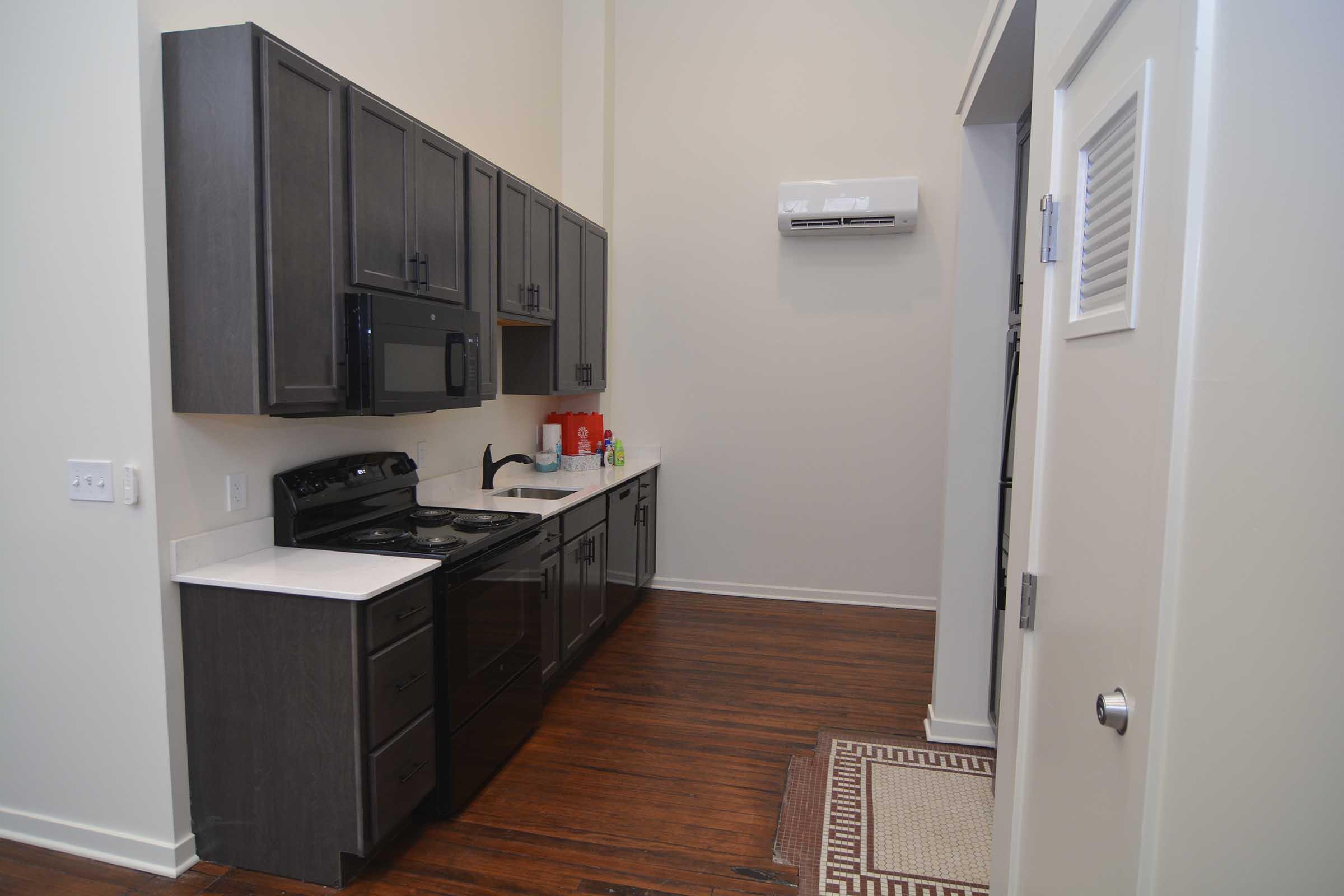 Modern kitchen with dark cabinetry, a black stove, microwave, and refrigerator. Light-colored countertops and wooden flooring provide a warm ambiance. An air conditioning unit is mounted on the wall, and a patterned rug is placed at the entrance. The space is tidy and well-lit, emphasizing a contemporary design.