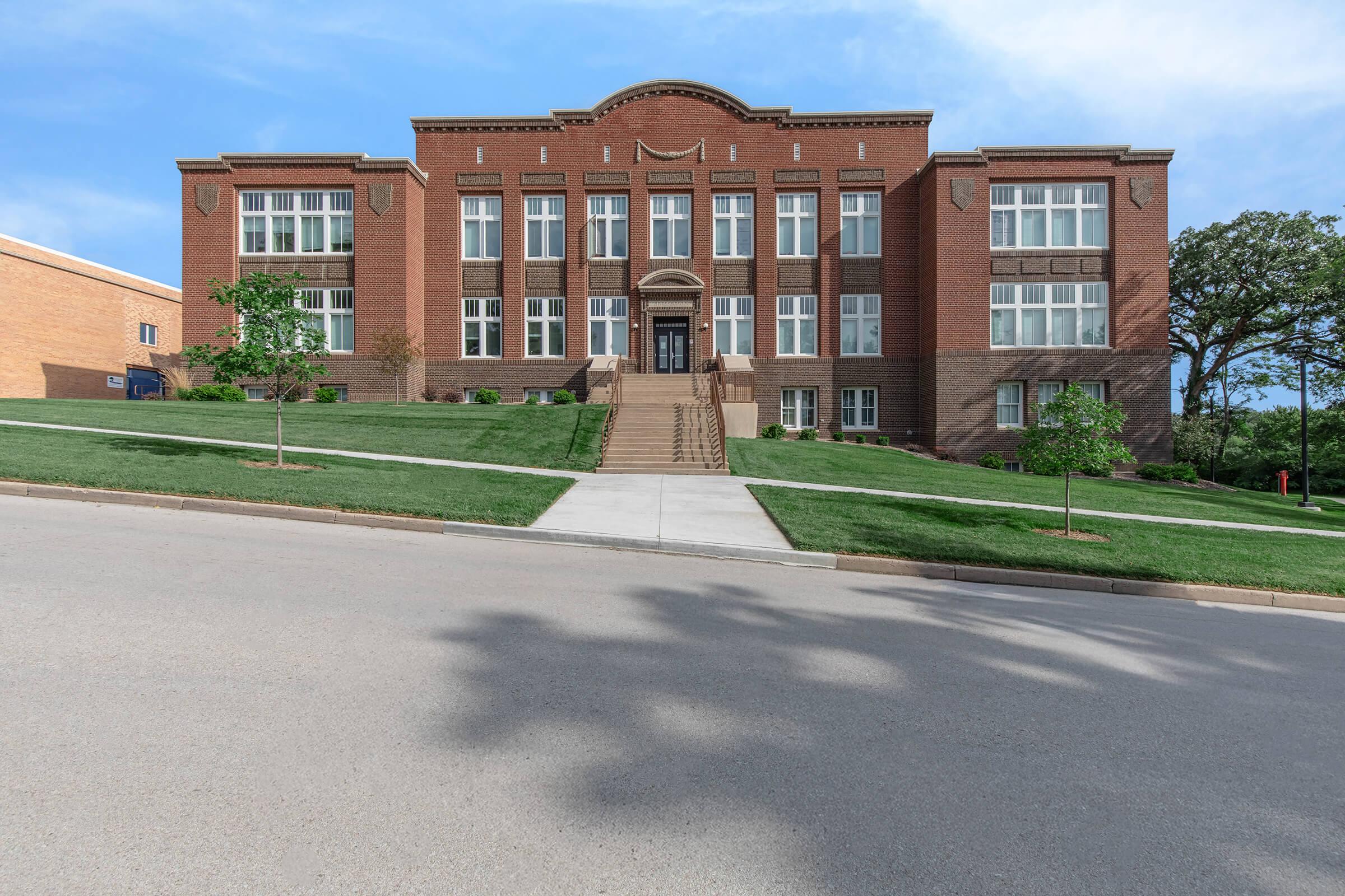 A large, two-story brick school building with a symmetrical facade, featuring tall windows and a prominent entrance with stairs. Surrounding the building is a landscaped lawn with small trees, and the image captures the building from a sloping street, under a clear blue sky.