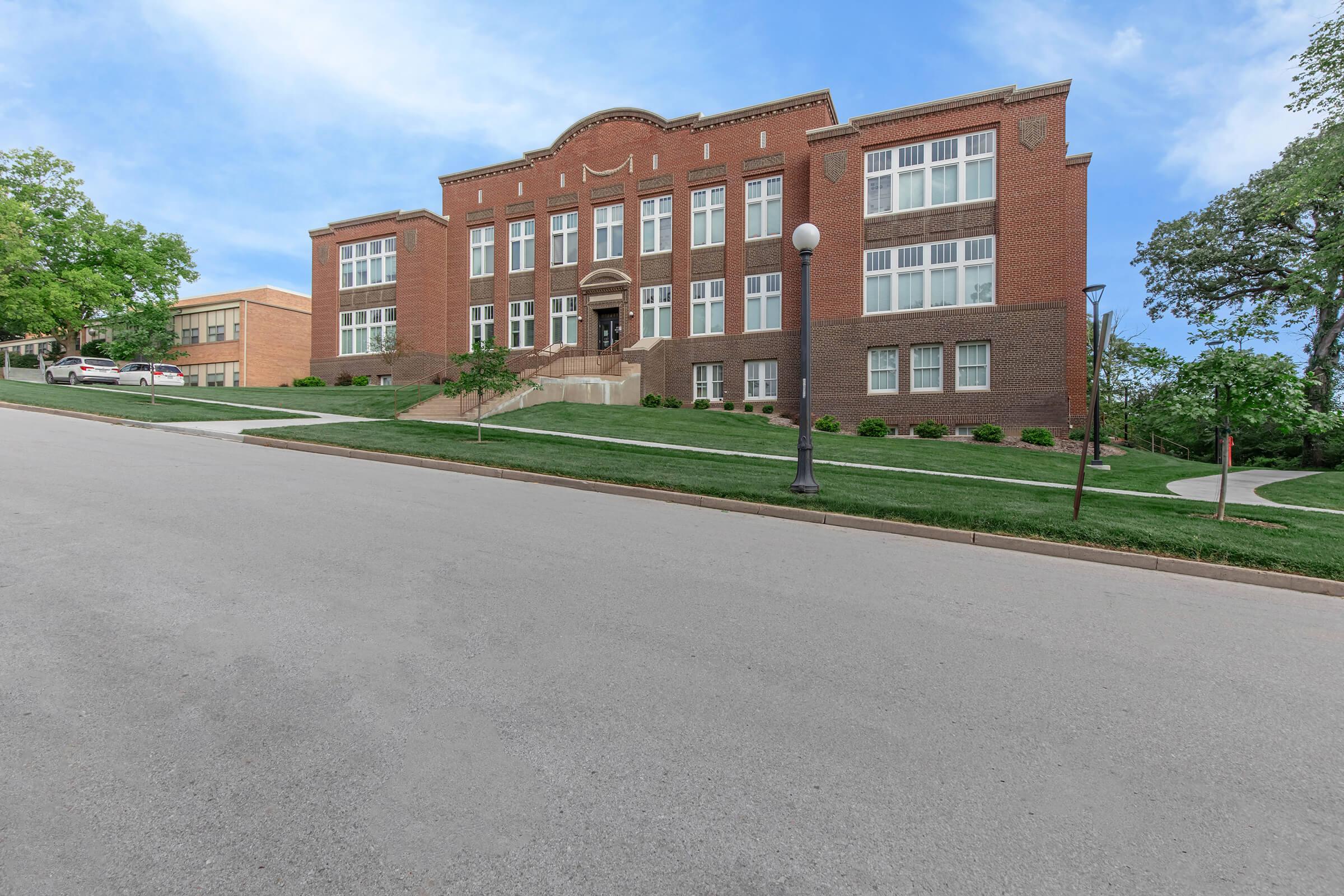 A large brick school building with multiple windows, situated on a sloped street. The landscape features green grass and small bushes, with a lamp post in front. In the background, there is another smaller building. The sky is clear with a hint of blue.