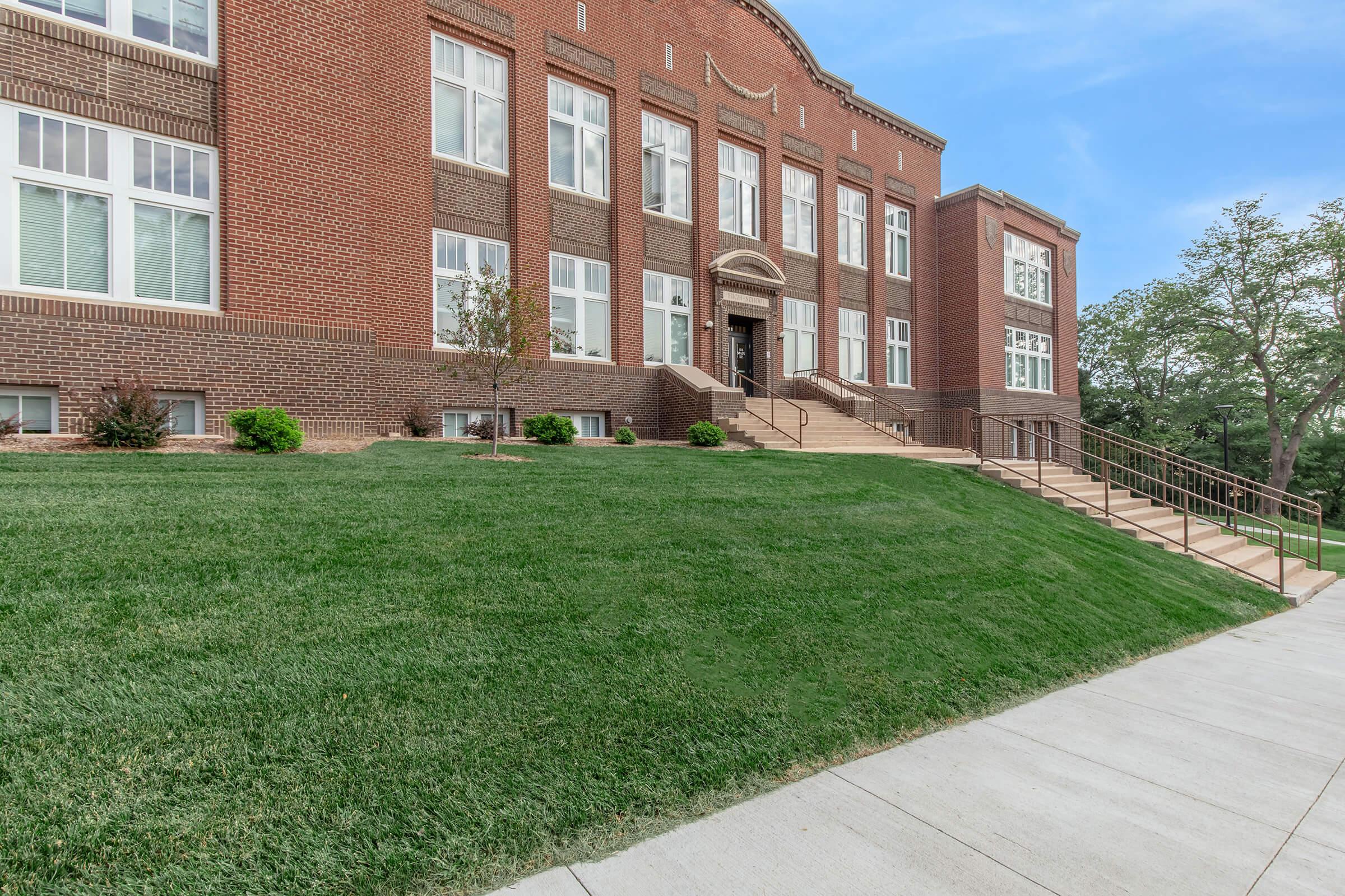 A brick building with large windows and a central entrance, surrounded by well-groomed grass and small trees. A concrete walkway leads to the entrance, and there are stairs leading up to it. The sky is clear and blue, suggesting a bright day.