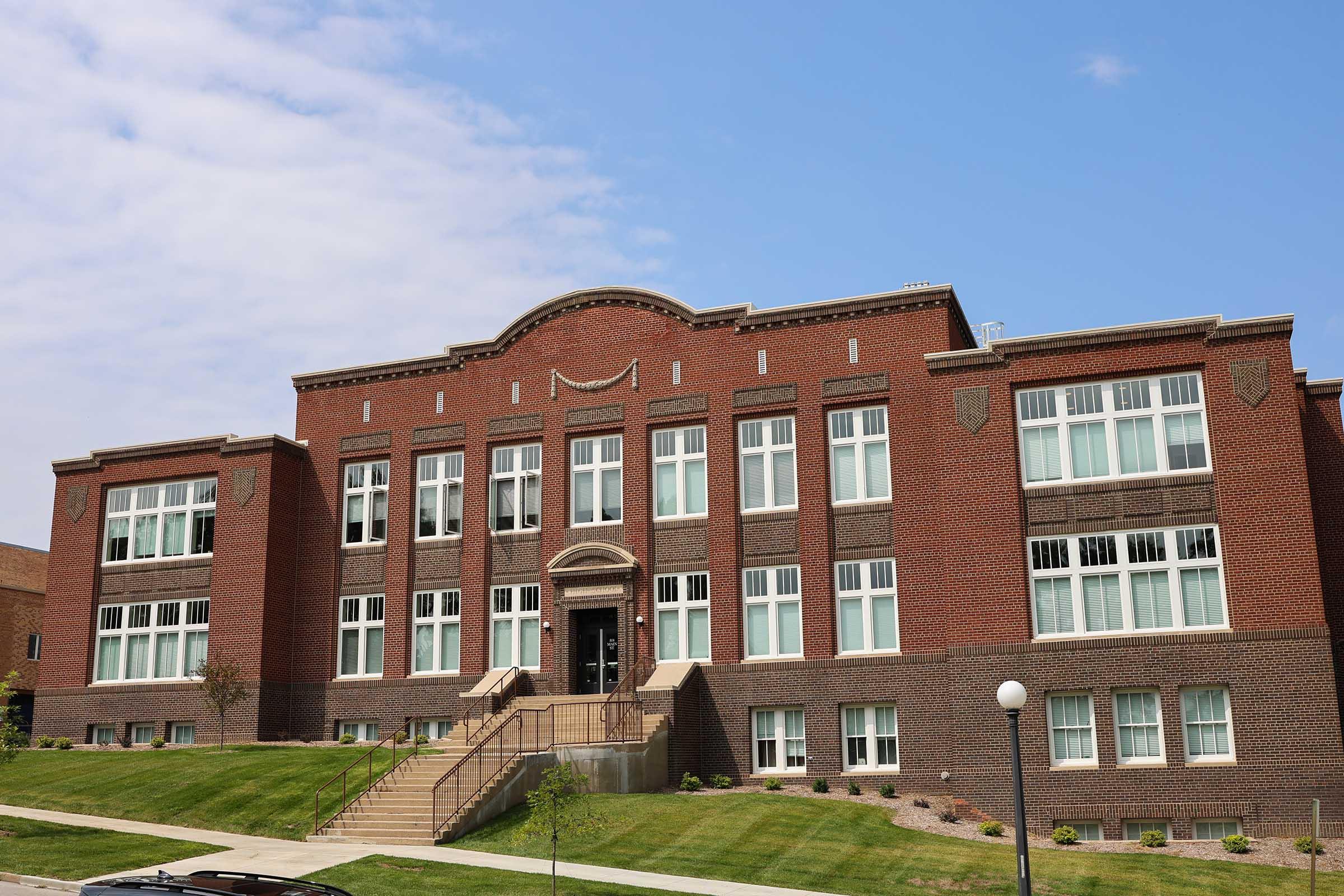 A historic brick building with large windows and a grand entrance, flanked by steps leading up to the front door. The architecture features decorative elements and a symmetrical design, set against a clear blue sky. Lush green landscaping surrounds the structure.