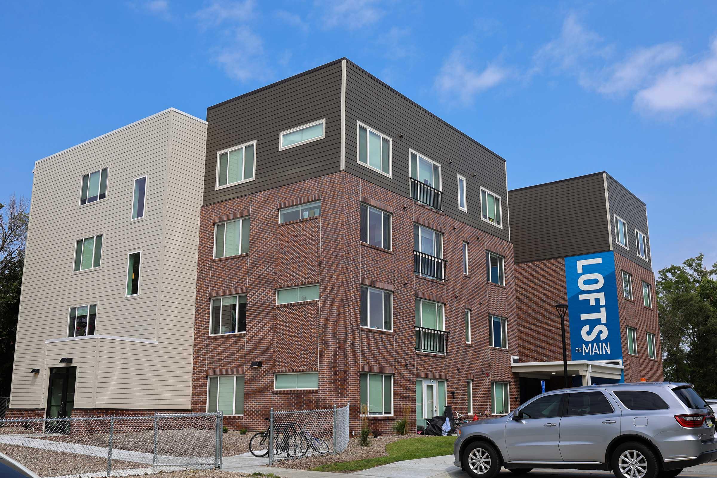 A modern brick apartment building with large windows, featuring a prominent sign that reads "Lofts on Main." The building has a mix of textures, including a cream-colored section. Bicycles are parked outside, and there is a silver SUV in the foreground against a clear blue sky.