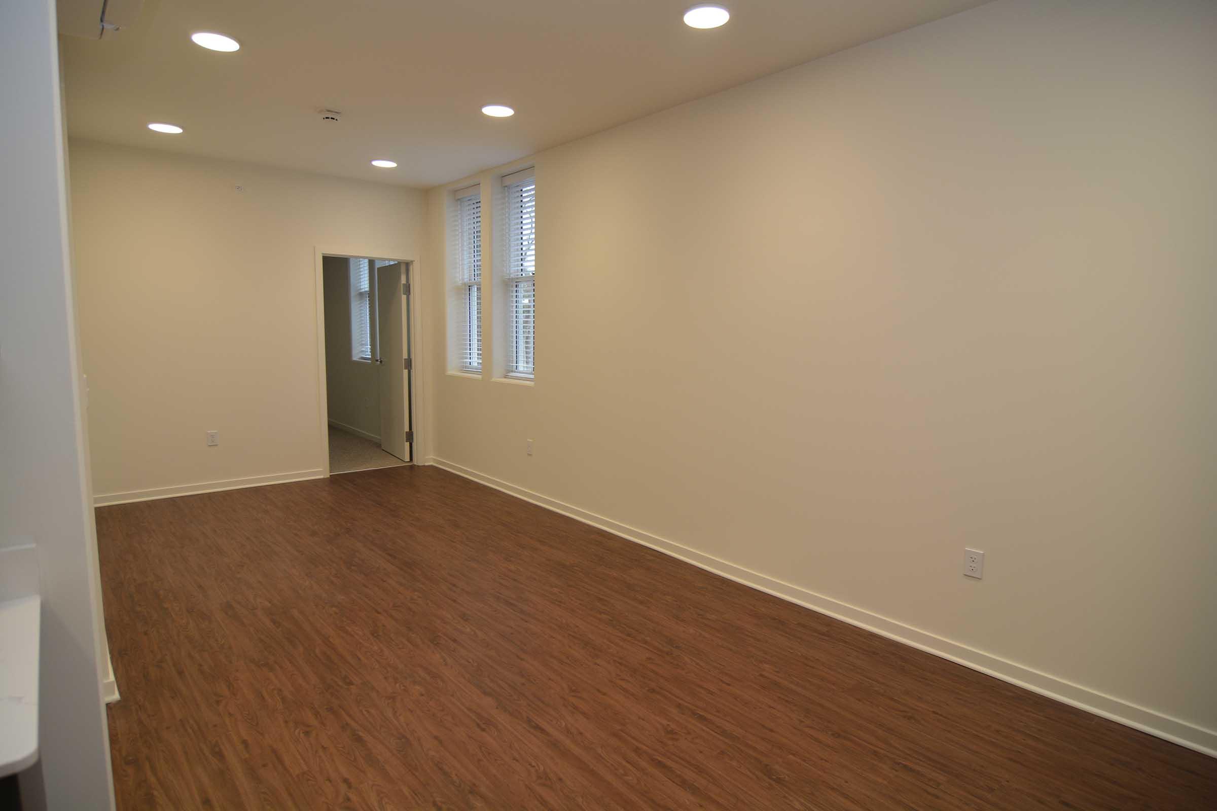 A spacious, empty room with light wood flooring and white walls. Recessed lighting illuminates the space, which features large windows on one wall. A doorway leads to another room, creating an open and airy atmosphere suitable for various uses.