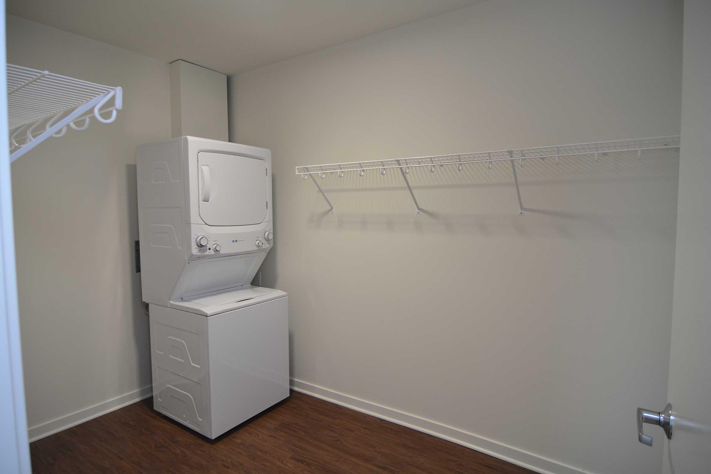 A small laundry room featuring a stacked washer and dryer setup against a light-colored wall. There is a wire shelf mounted above, providing storage space, and the floor is finished in a wooden texture. The overall space is clean and uncluttered, with neutral tones.