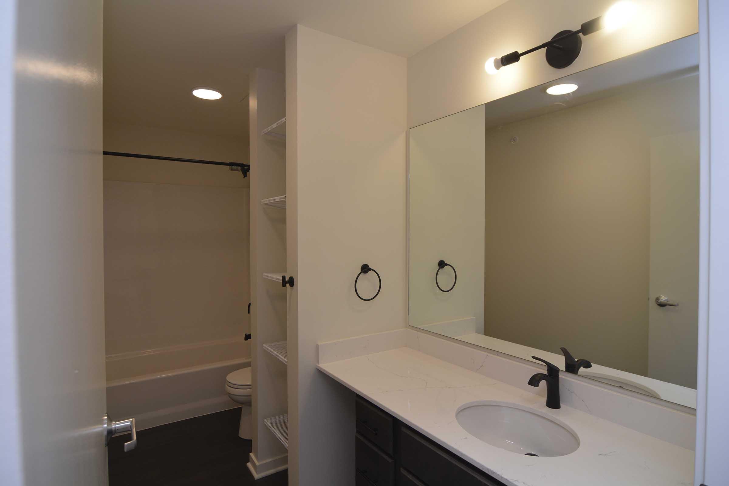A modern bathroom featuring a light-colored wall, a white countertop with a sink and black faucet, a large mirror, and shelving. The space includes a bathtub with a shower rod, a toilet, and a towel ring. Bright lighting is provided by wall-mounted fixtures. The floor is dark, contrasting with the lighter walls.