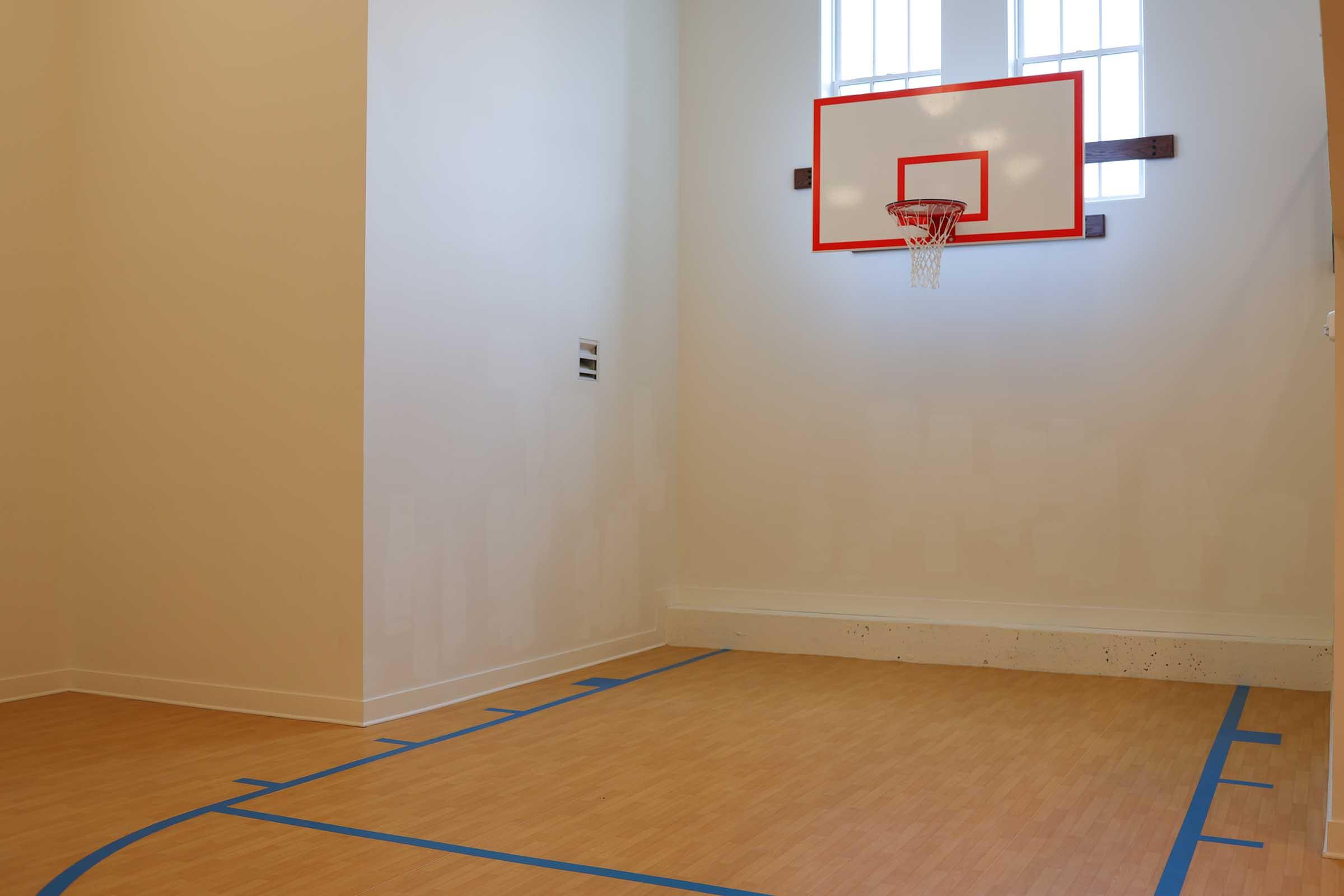 Indoor basketball court with a wooden floor, featuring a wall-mounted basketball hoop outlined in red. The space is minimally decorated, with large windows allowing natural light. Blue markings on the floor indicate the court boundaries.
