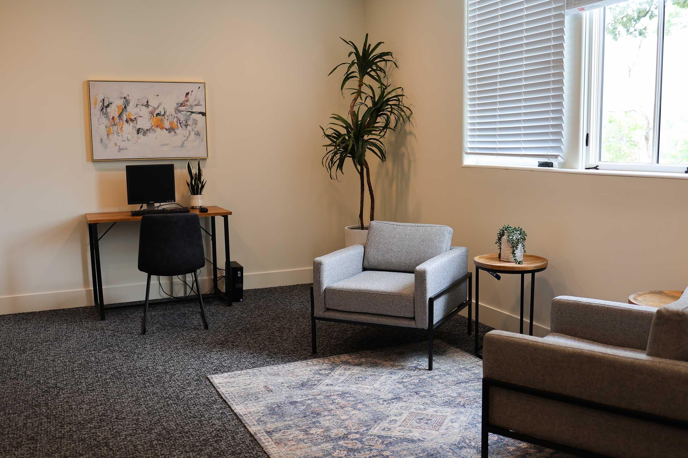 A modern, minimalist room featuring two gray armchairs, a small side table with a plant, a desk with a computer and a decorative plant, and a large window with blinds. The floor is covered with carpet, and there’s a colorful abstract painting on the wall.
