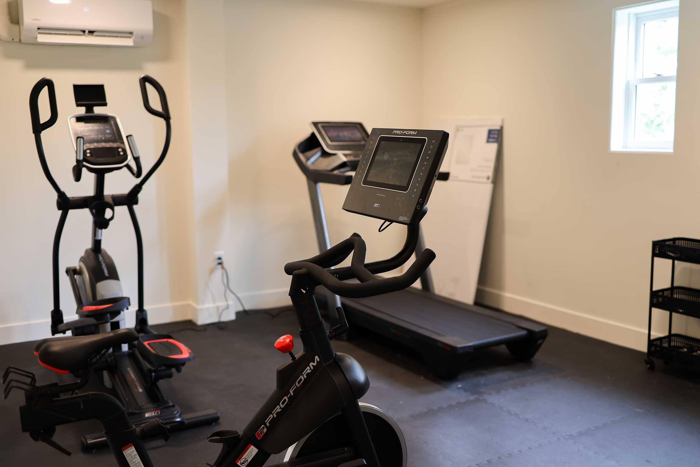 A small home gym featuring a stationary bike and an elliptical machine in the foreground, with a treadmill in the background. The room has light-colored walls and a window allowing natural light, along with an air conditioning unit mounted high on the wall.
