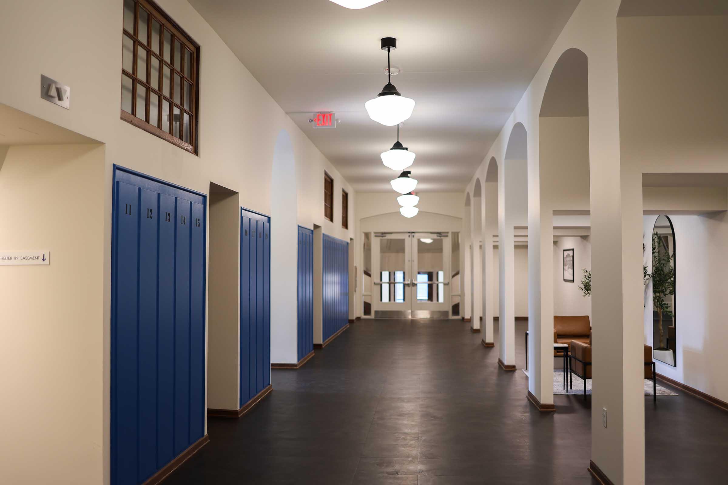 A spacious, well-lit hallway featuring blue lockers on one side, arched doorways, and decorative light fixtures. The floor is dark, and there are chairs positioned near a large window at the far end, creating a welcoming atmosphere in a school or institutional setting.