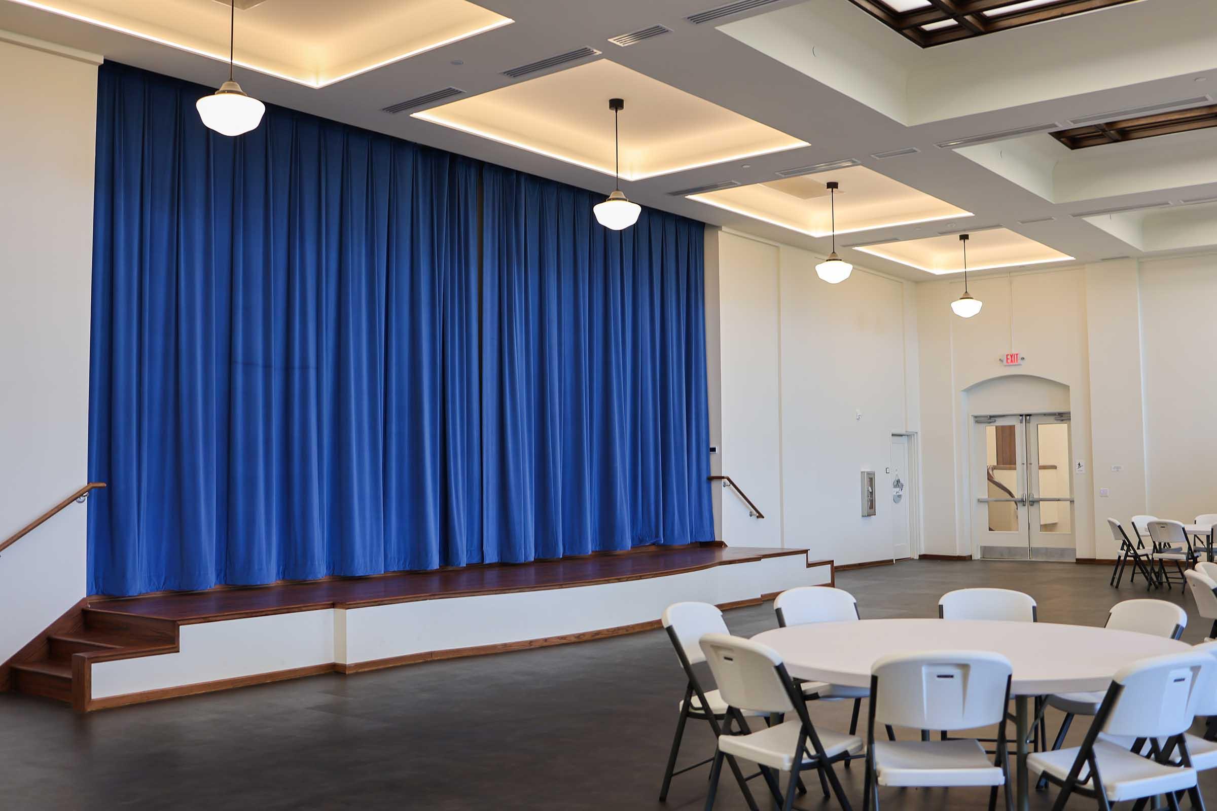 A spacious room with a blue curtain backdrop stage elevated at one end. The area features several round tables and chairs arranged for seating. Soft lighting from pendant lamps illuminates the space, while large windows provide natural light.