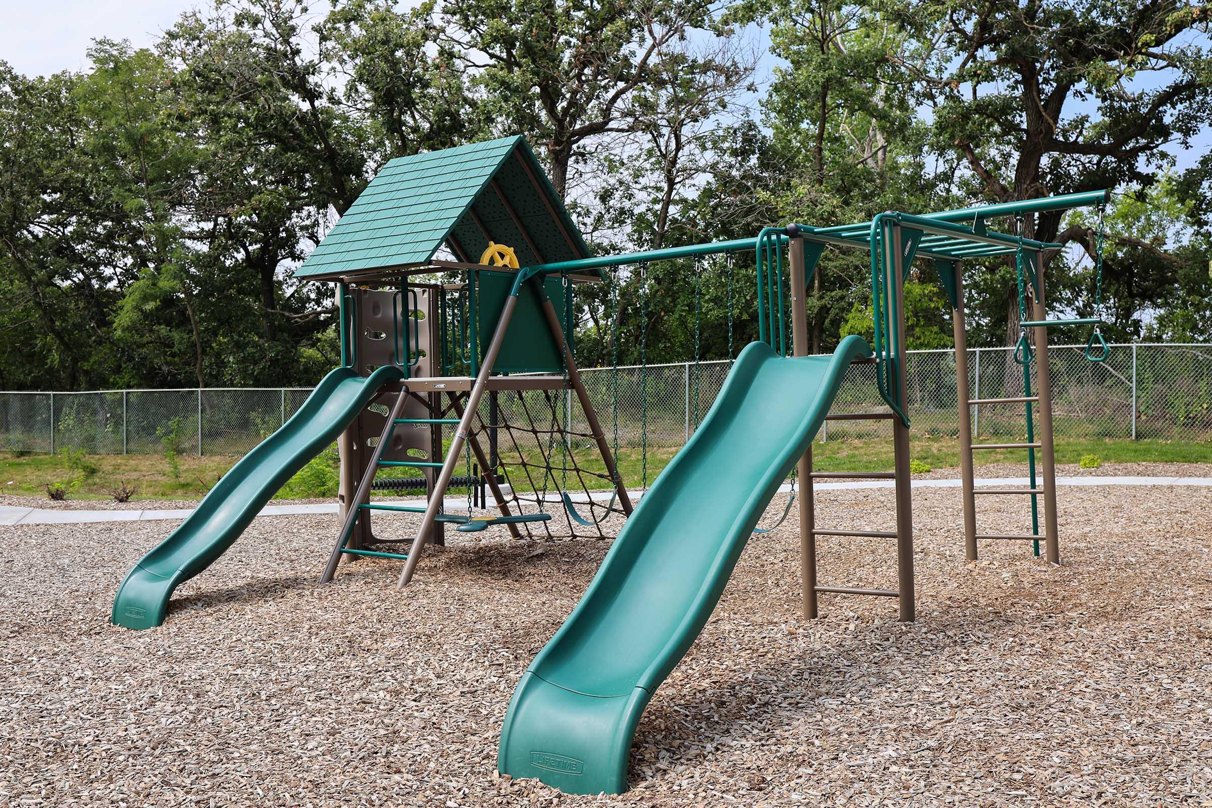 A playground structure featuring two green slides, a climbing frame, and a playhouse with a sloped roof. The playground is surrounded by a gravel area and backed by trees, offering a natural setting for children to play.