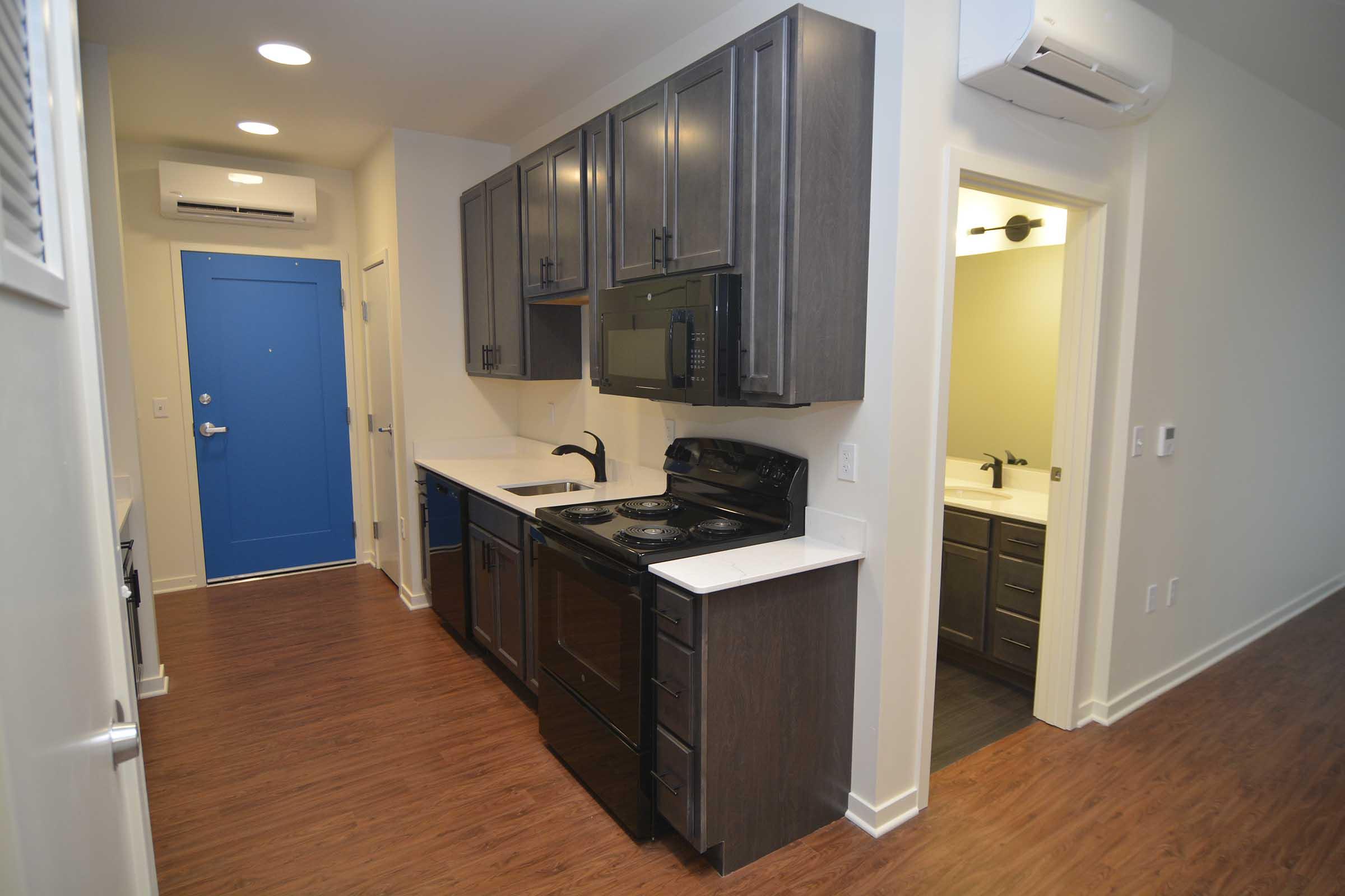 A modern kitchen with dark wood cabinets, a black stove, microwave, and refrigerator. White countertops and light-colored walls enhance the bright space. An air conditioning unit is visible, along with an entrance door painted blue. A doorway leads to a bathroom area. Wooden flooring completes the contemporary look.