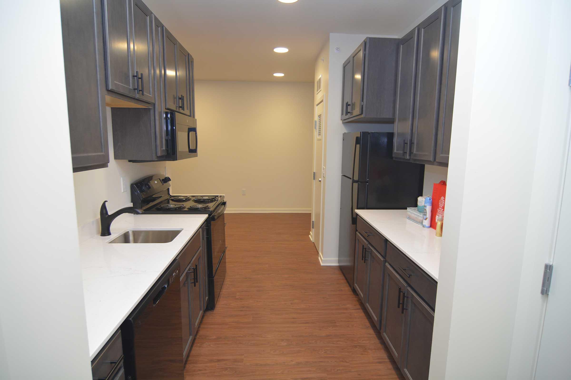 A modern kitchen with dark wood cabinets, stainless steel appliances including a stove and refrigerator, white countertops, and a sink. The layout features a narrow passage with a view of a well-lit space at the end. The flooring is a warm-colored laminate, creating a cozy atmosphere.