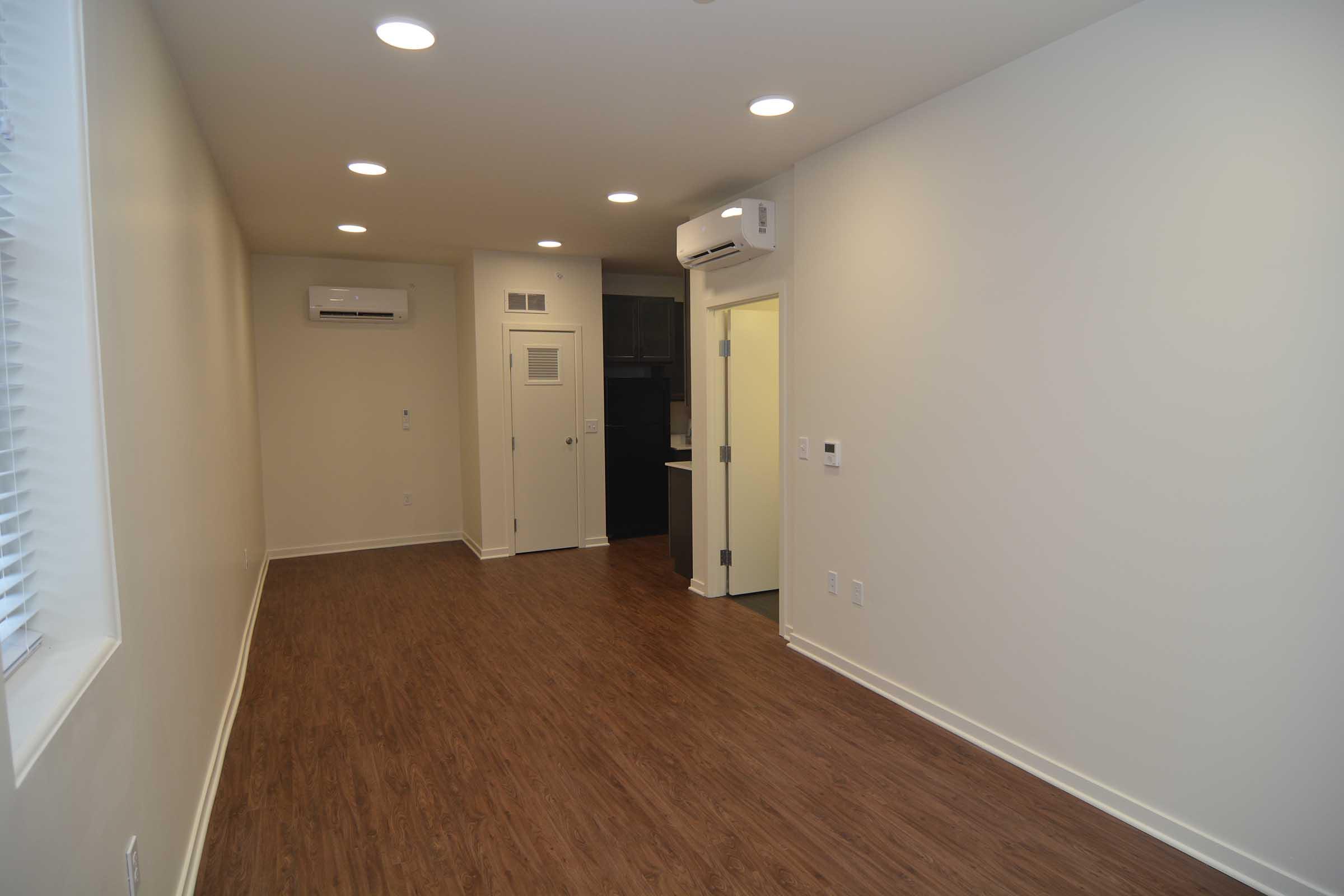 A minimalist interior of a room featuring light-colored walls, hardwood flooring, and recessed lighting. It has a doorway leading to a kitchen area in the background, an air conditioning unit mounted on the wall, and windows with blinds allowing natural light.