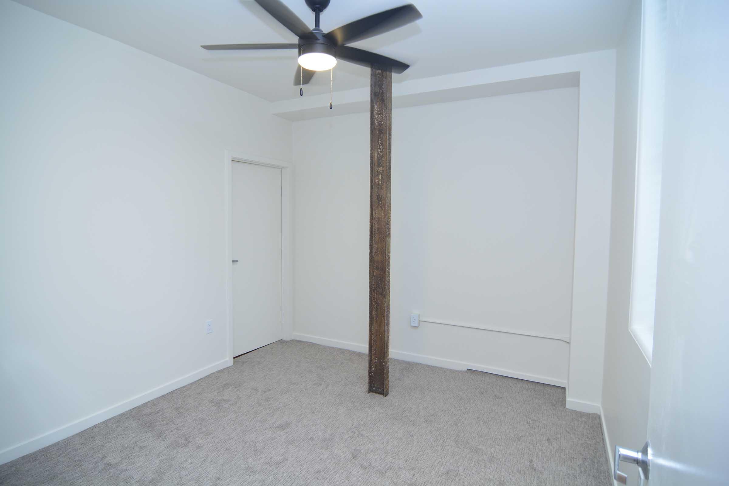 A sparsely furnished room featuring light-colored walls and carpeted flooring. There is a ceiling fan with black blades and a wooden support beam in the center. A closed white door is visible on one wall, and there is a small window allowing natural light in. The overall aesthetic is minimalistic and modern.