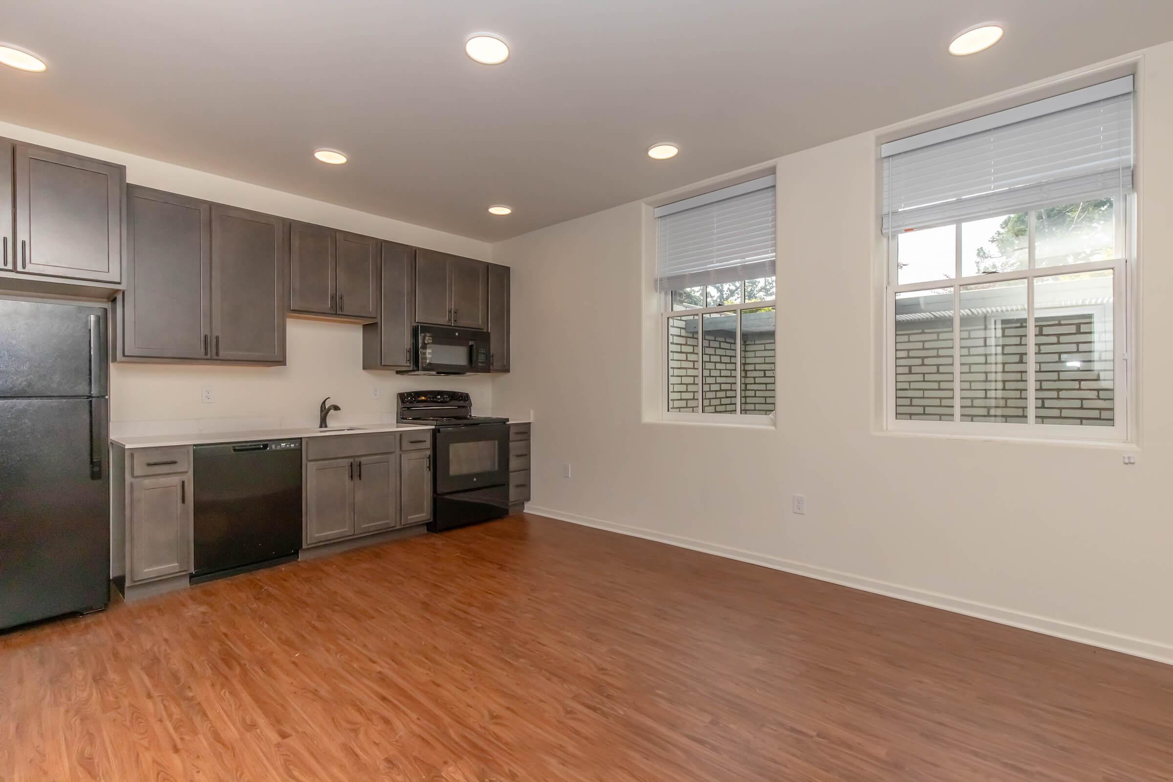 Modern kitchen in a living space featuring dark cabinetry, a black refrigerator, and stainless steel appliances. The floor is a clean, light wood, and there are two windows allowing natural light into the room. The walls are painted in a neutral tone, creating a bright and inviting atmosphere.