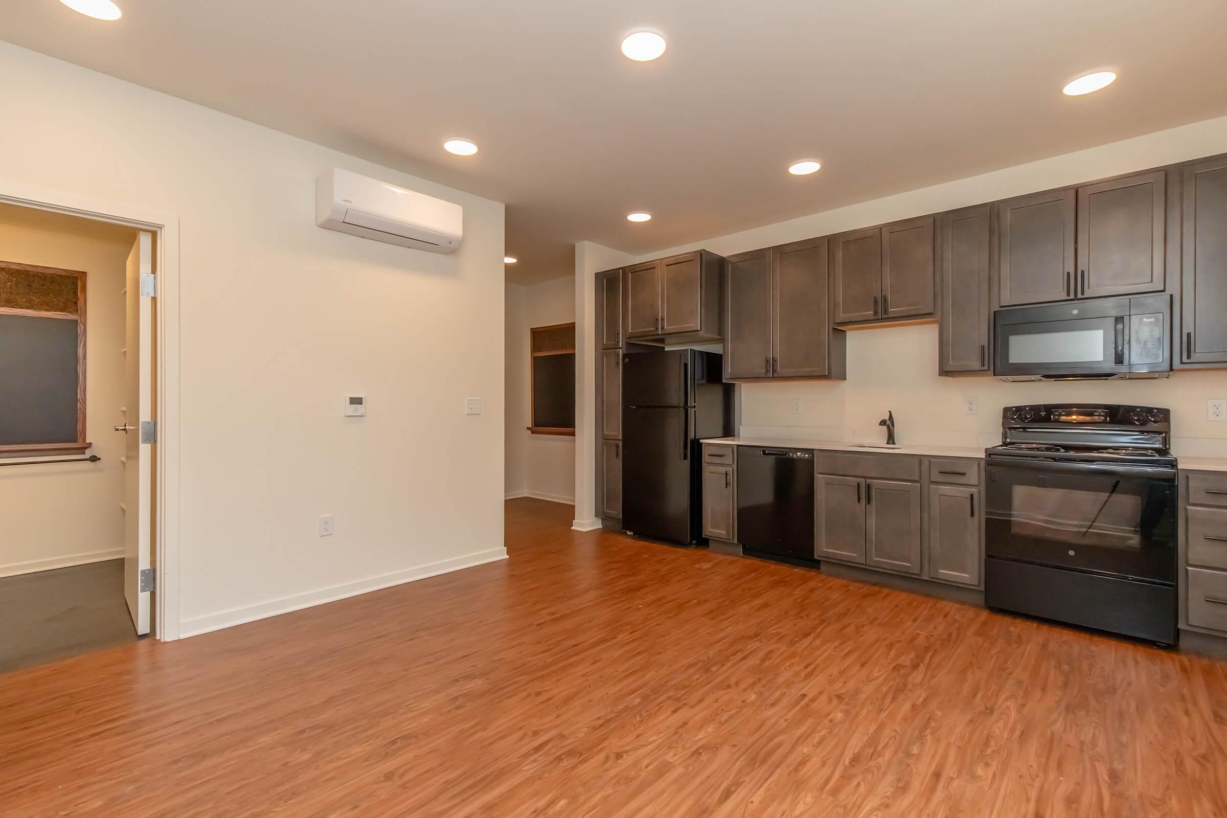 Modern kitchen with dark cabinetry, stainless steel appliances, and a wooden floor. Features include a black refrigerator, stove, and a window above the sink, providing natural light. The space is open and well-lit, with air conditioning and a door to another room visible.