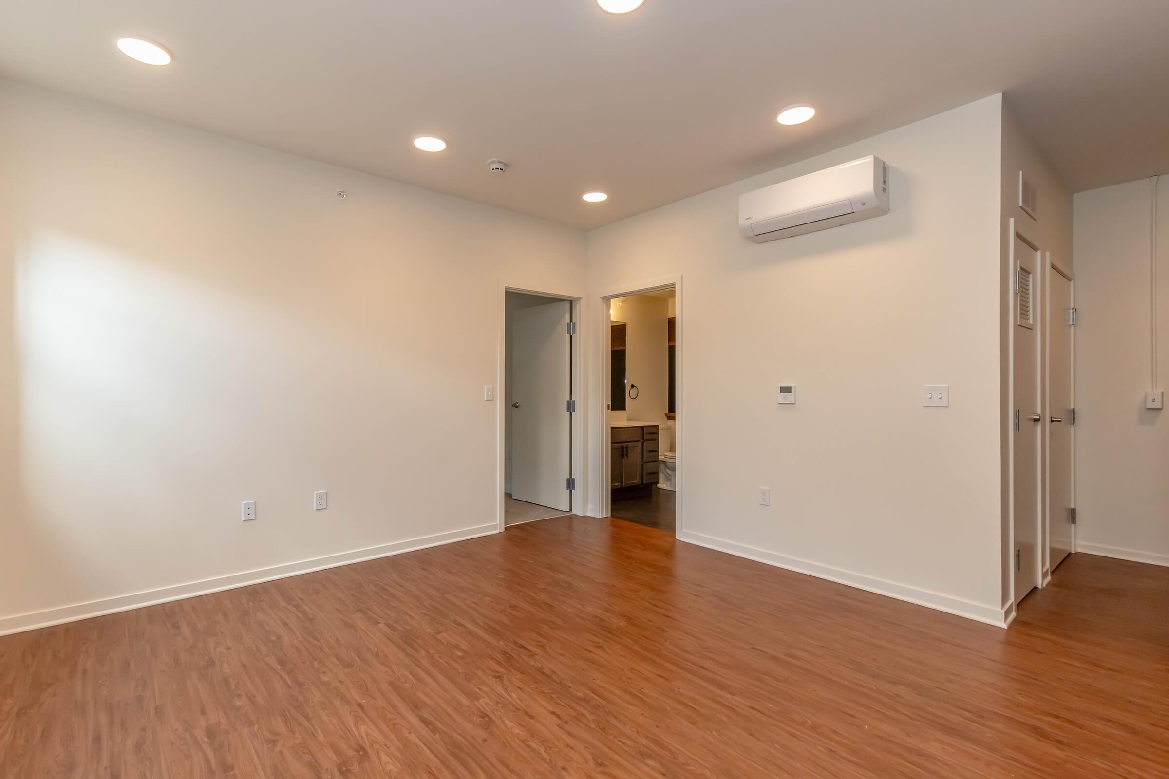Empty room with light-colored walls and a wooden floor, featuring recessed lighting in the ceiling. There is an open doorway leading to a bathroom, a wall-mounted air conditioning unit, and a control panel on the wall. The space is bright and minimalistic, showing no furniture or decor.