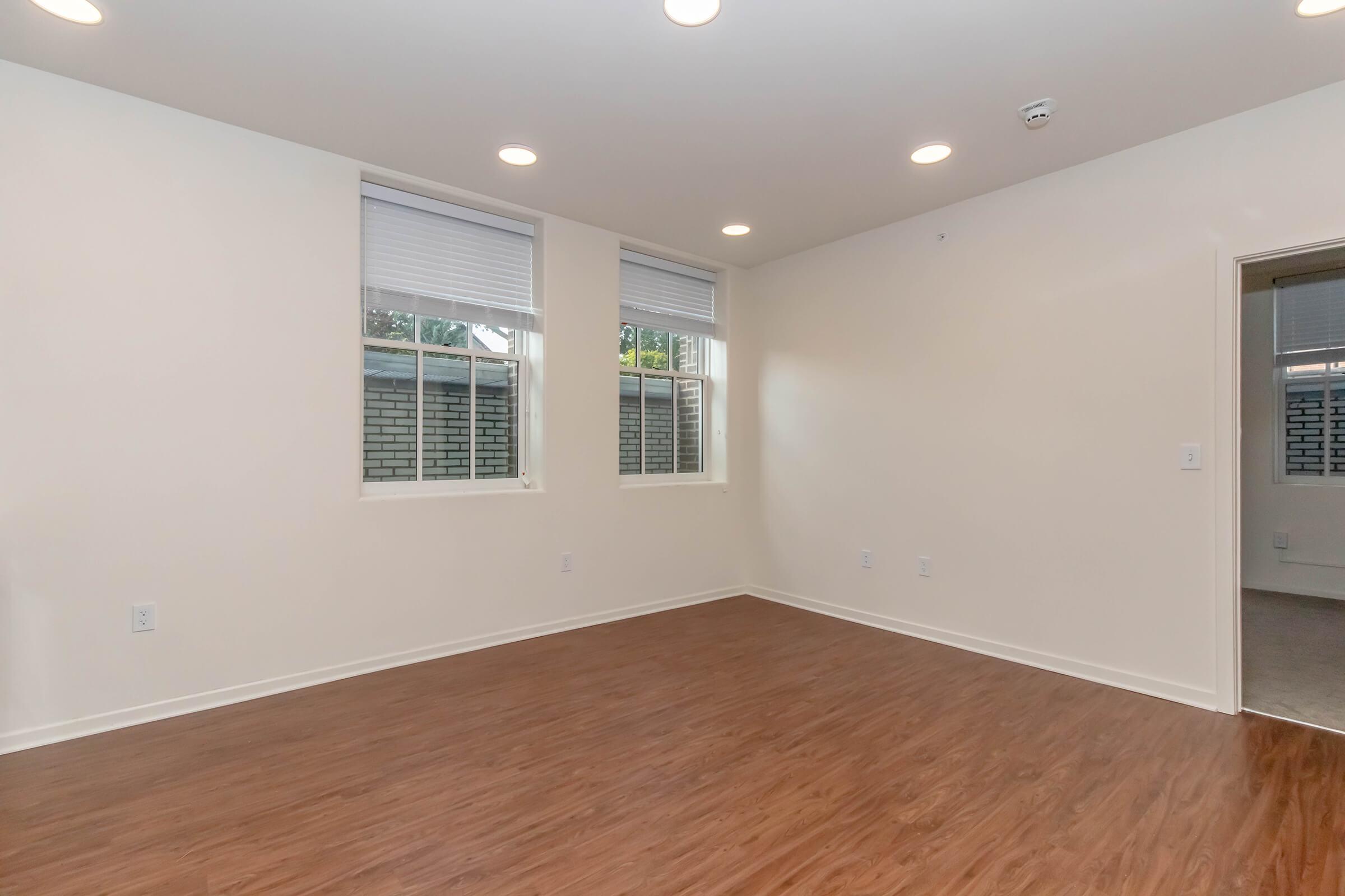 A bright, empty room featuring light-colored walls and hardwood flooring. There are two windows with white blinds, allowing natural light to enter. The space is uncluttered, emphasizing its size and potential for various layouts and decorations.