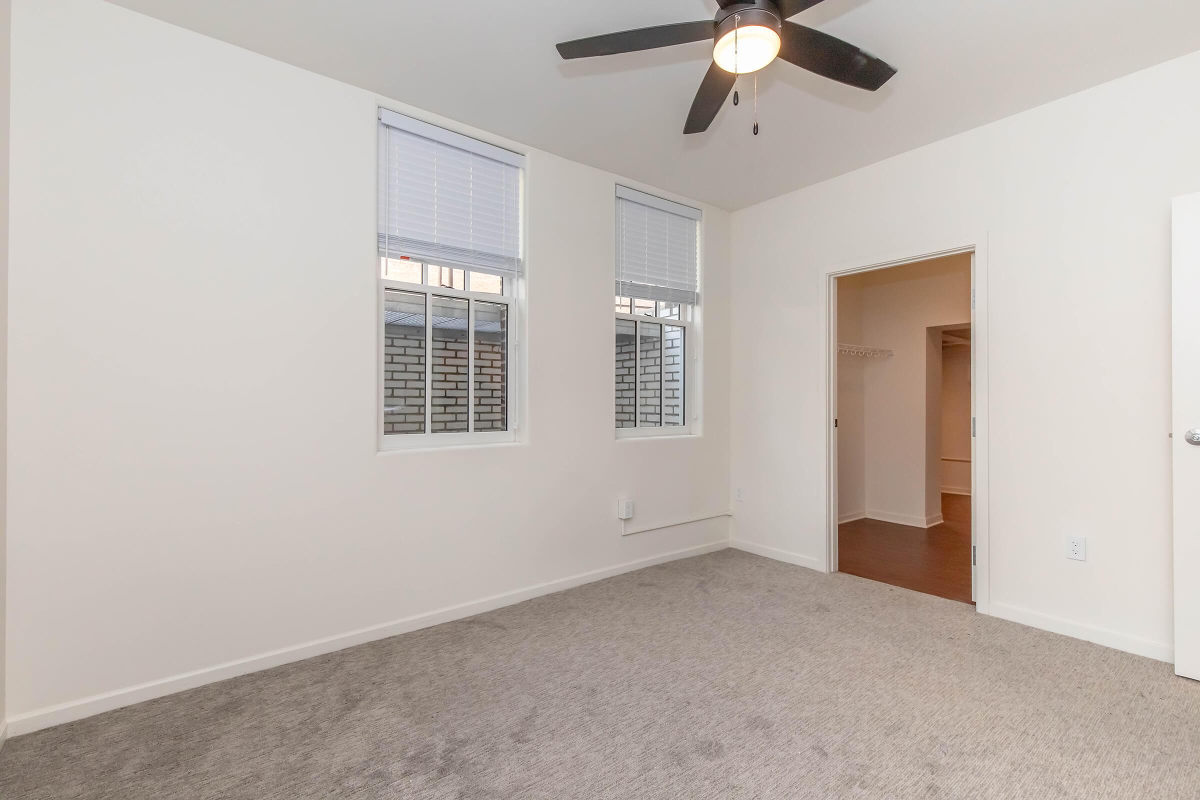 A well-lit room with light gray walls and carpet. There are two windows with white blinds allowing natural light. A ceiling fan is mounted above. On one side, there is an open door leading to another area, while the opposite side remains blank, creating a spacious, clean appearance.