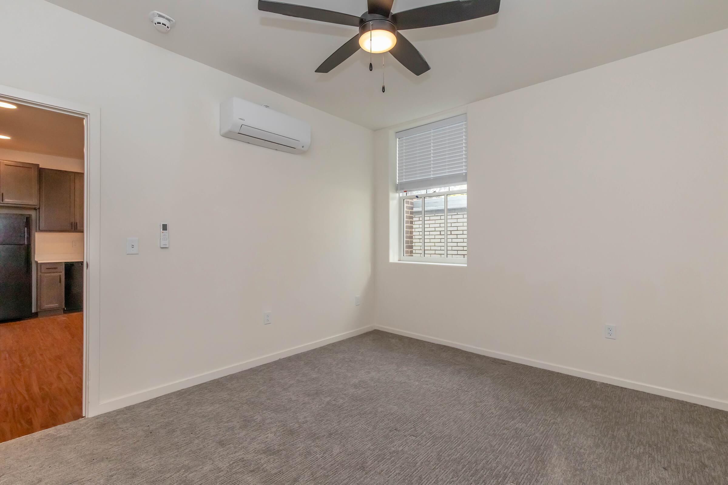 A small, empty room with light-colored walls and carpeted flooring. A ceiling fan hangs in the center, and there is a window on one wall letting in natural light. A doorway leads to another area, possibly a kitchen or hallway, where dark cabinets are visible. The space appears modern and uncluttered.