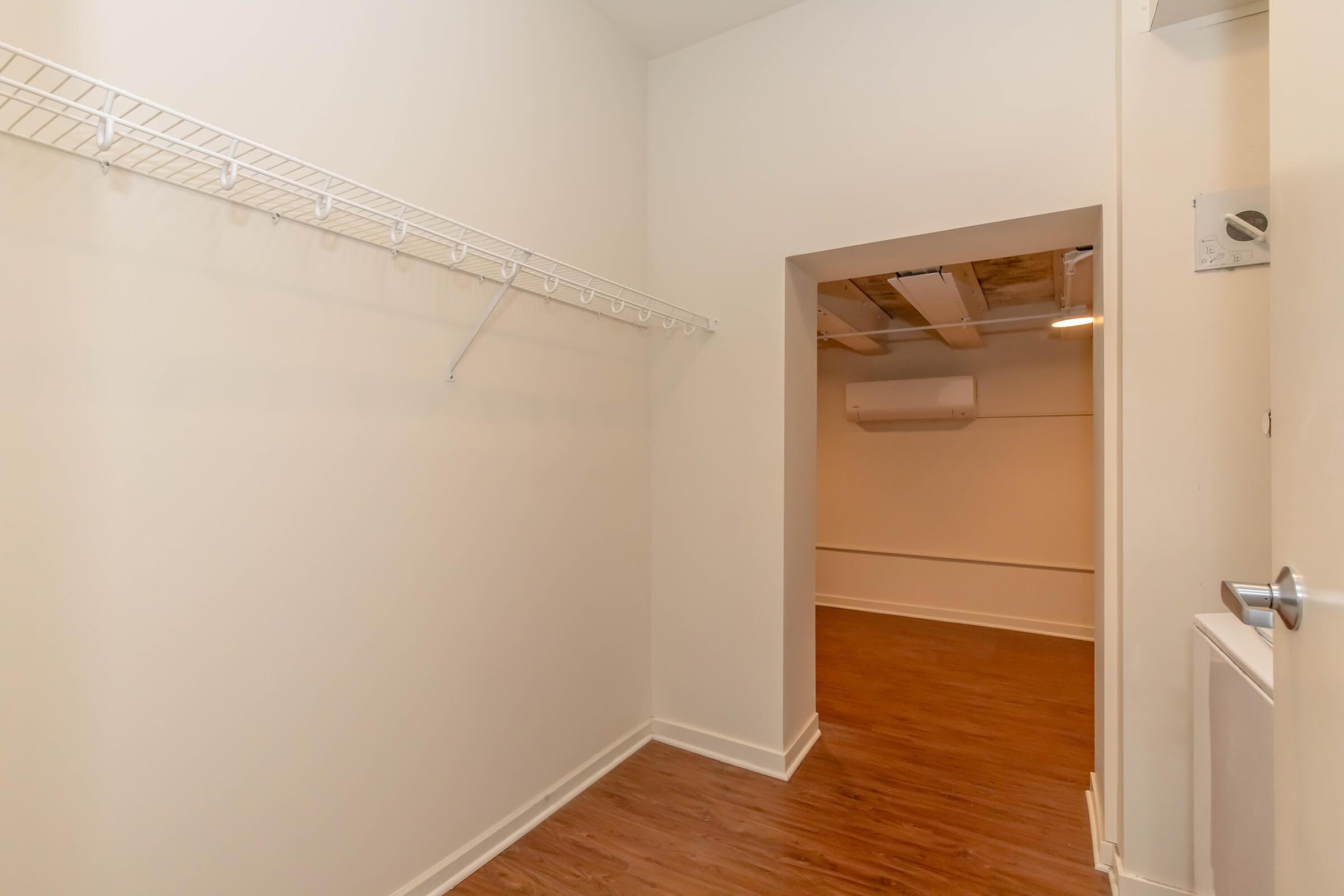 A spacious, empty closet with a wire shelf on the left side, featuring white walls and wooden flooring. An opening leads to a well-lit room in the background, revealing a modern, minimalistic design. The space appears tidy and uncluttered, suitable for storage or organization.