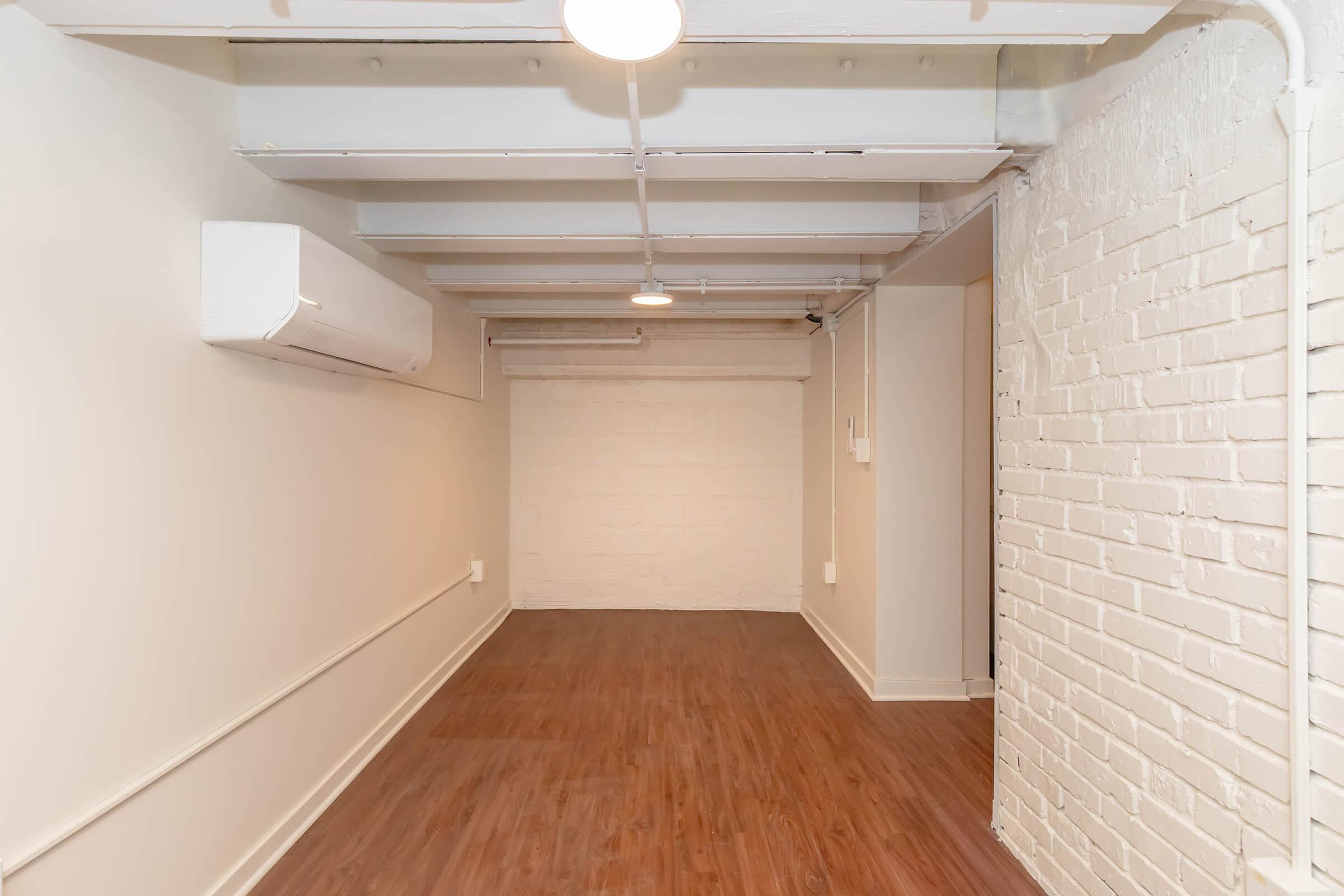 A spacious, unfurnished room with a wooden floor, white brick walls, and exposed beams. There is an air conditioning unit mounted on the wall. The area is well-lit with ceiling lights, creating a clean and minimalistic look, suitable for various purposes such as storage or office space.